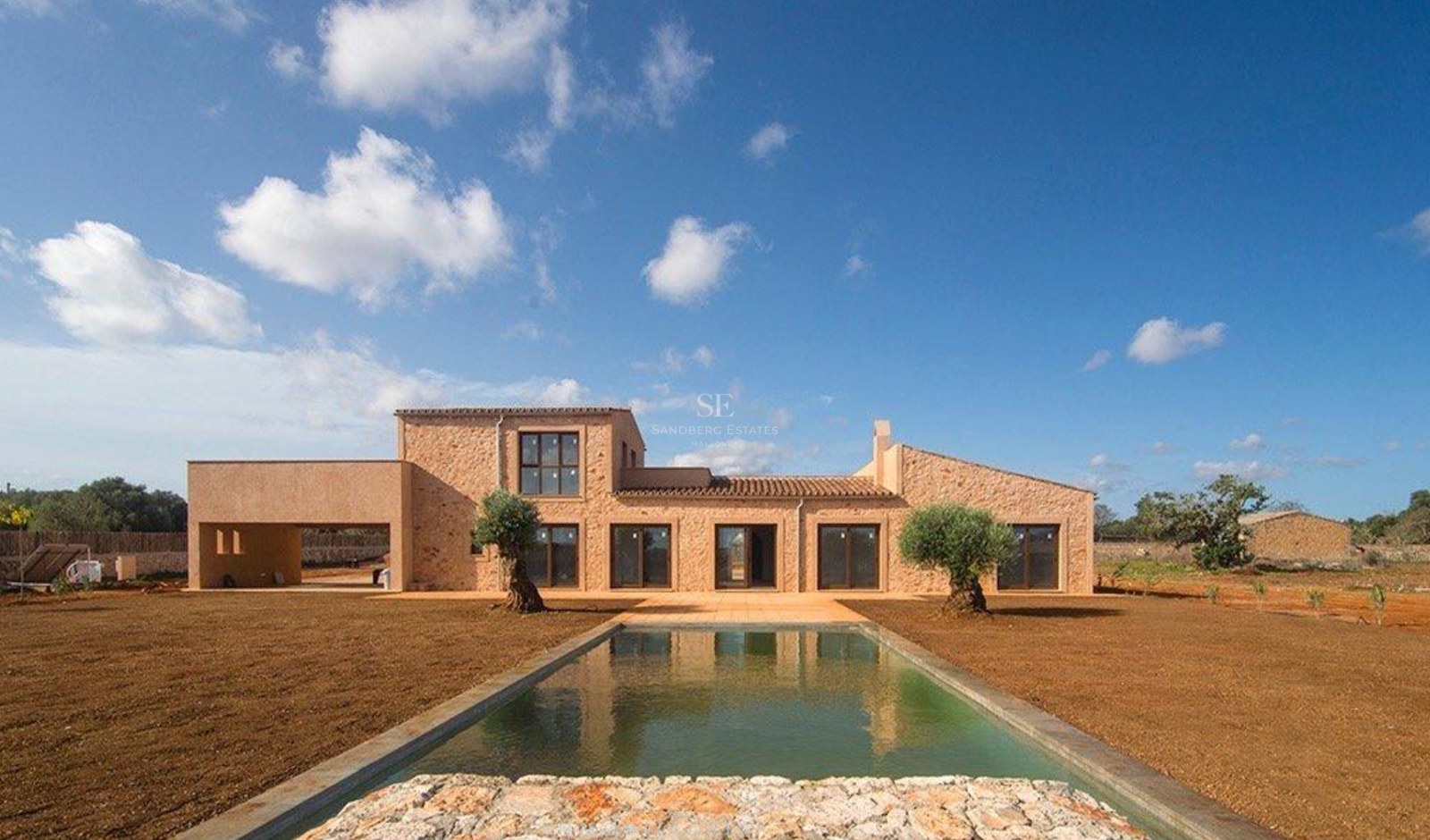 Exterior view of a natural stone villa featuring a rectangular swimming pool, olive trees, and large windows.