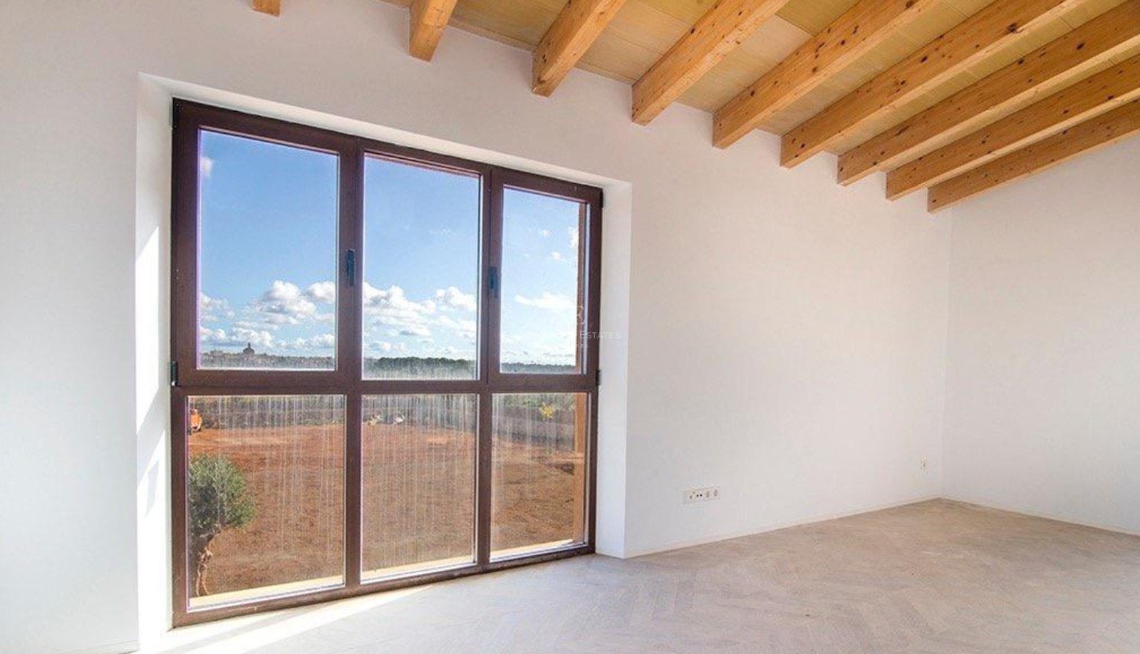Empty room featuring exposed wooden ceiling beams and a large dark-framed window overlooking a sunny rural plot.