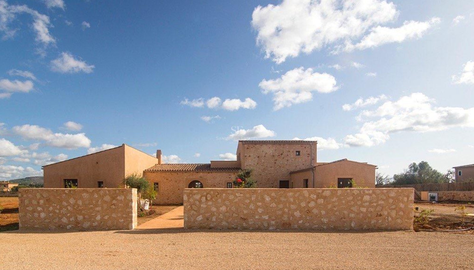 Exterior view of a stone and ochre finca under a blue sky, featuring a traditional stone wall and gravel entrance.