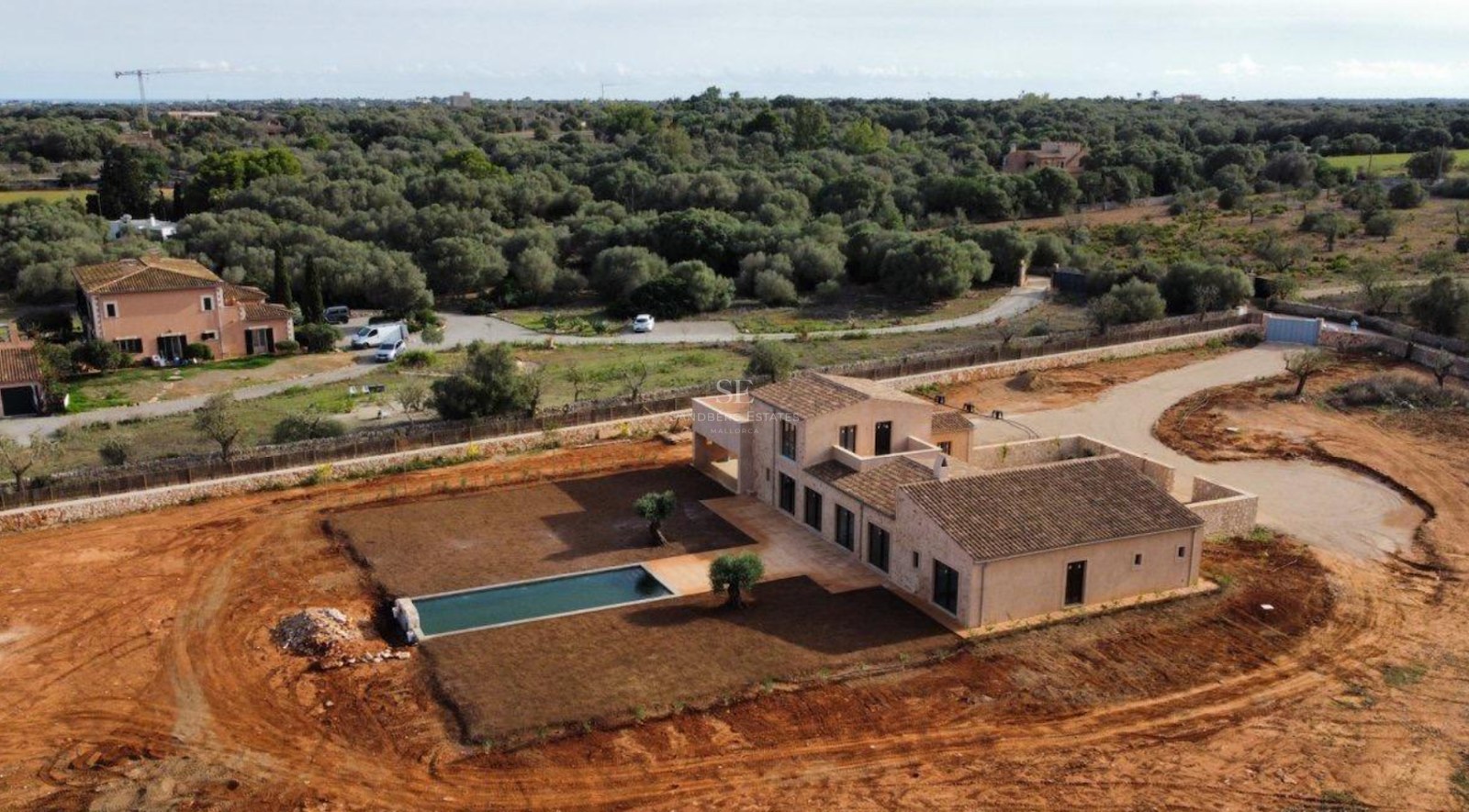 Aerial view of a newly built stone villa with a long swimming pool surrounded by a large plot and lush trees.