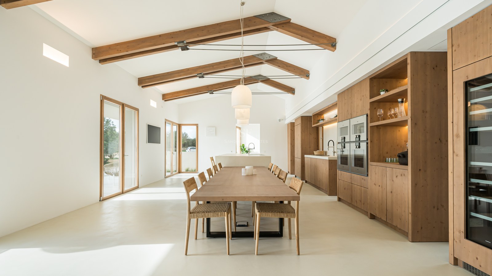 Bright open-plan dining room with vaulted ceilings, exposed wooden beams, and a large oak table next to a modern kitchen.