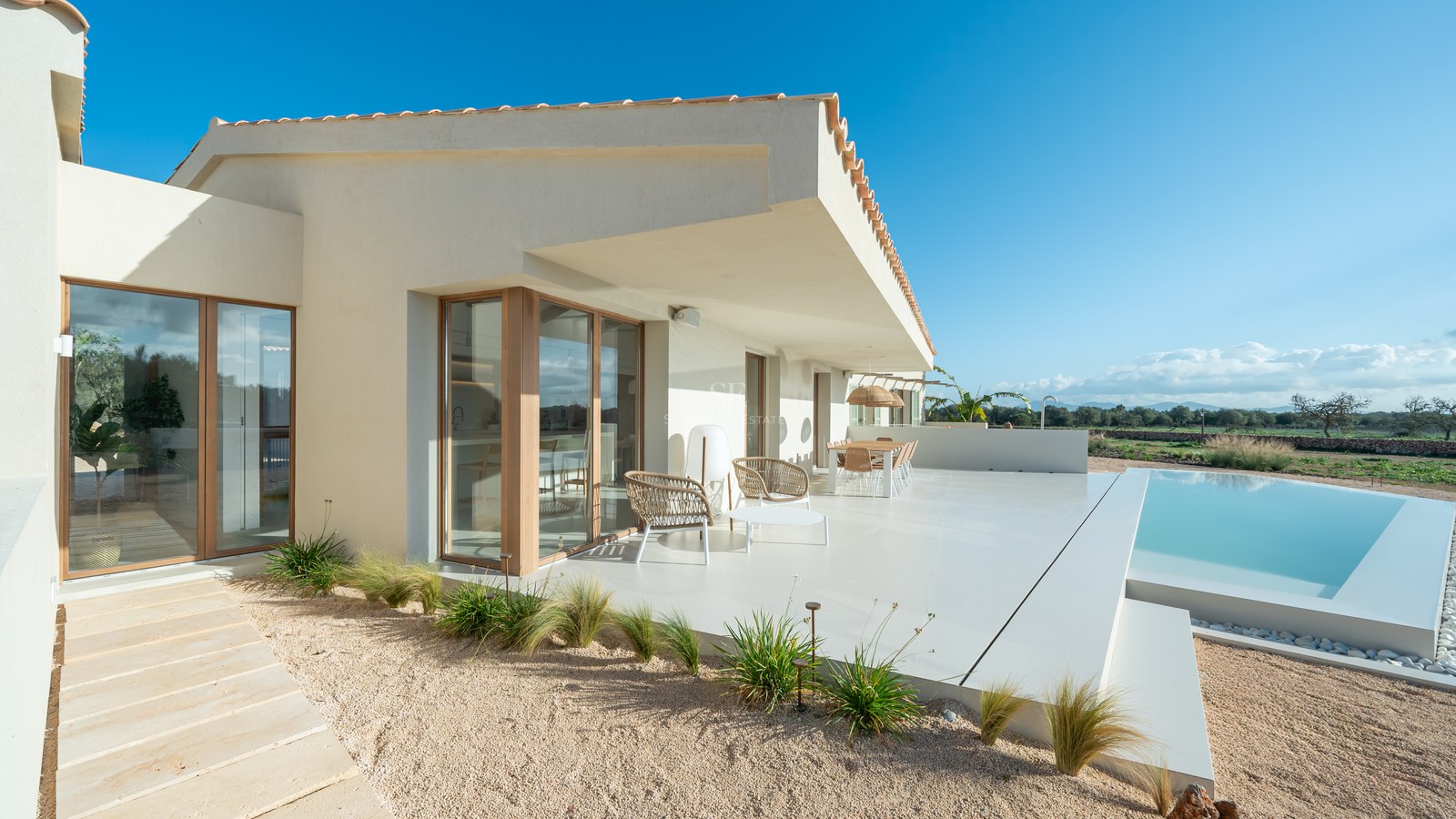 Modern villa terrace with white lounge furniture, wooden framed glass doors, and a turquoise infinity pool under clear sky.