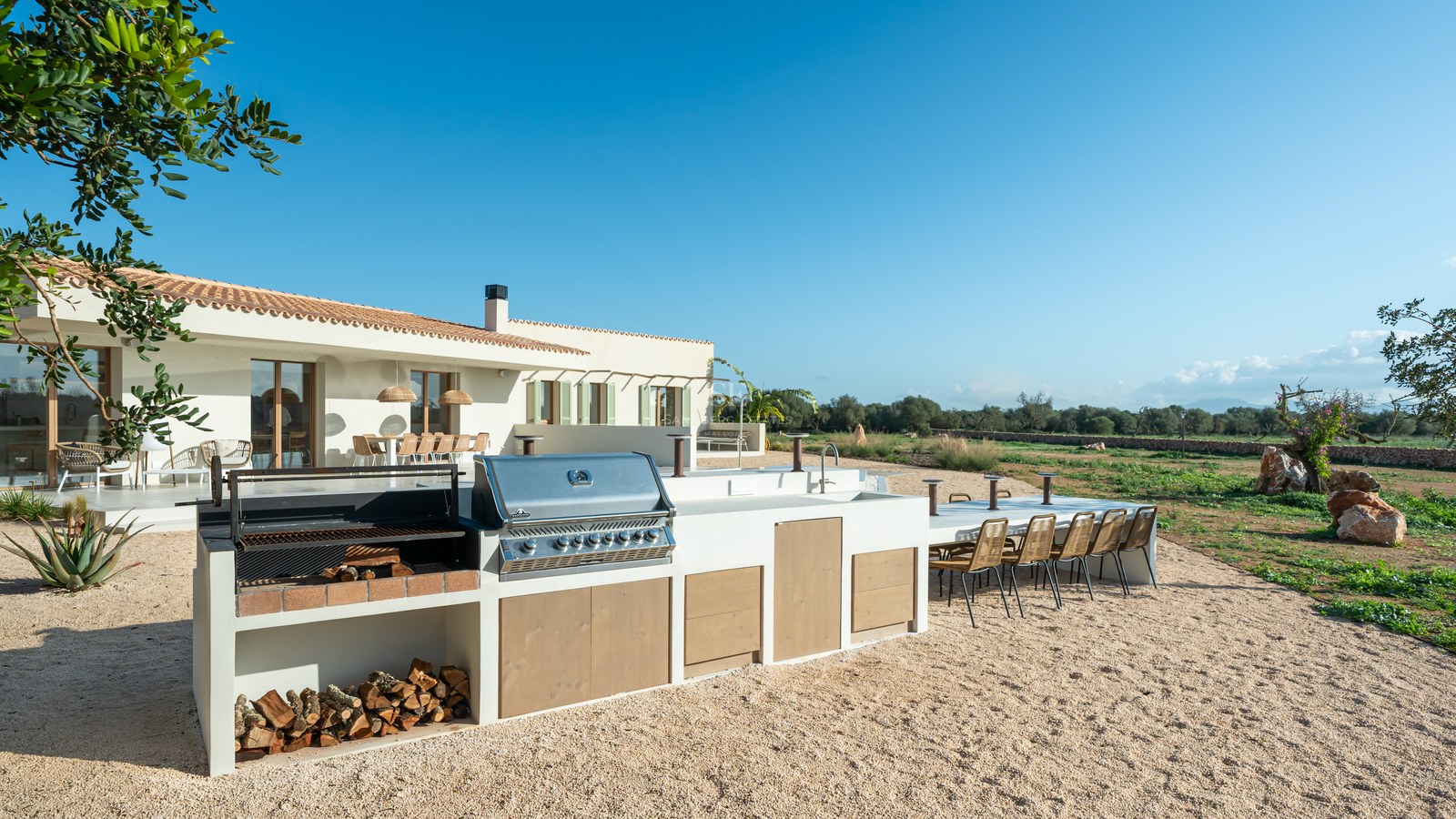 Modern outdoor kitchen with stainless steel grill, wood storage, and large dining table on a gravel terrace.