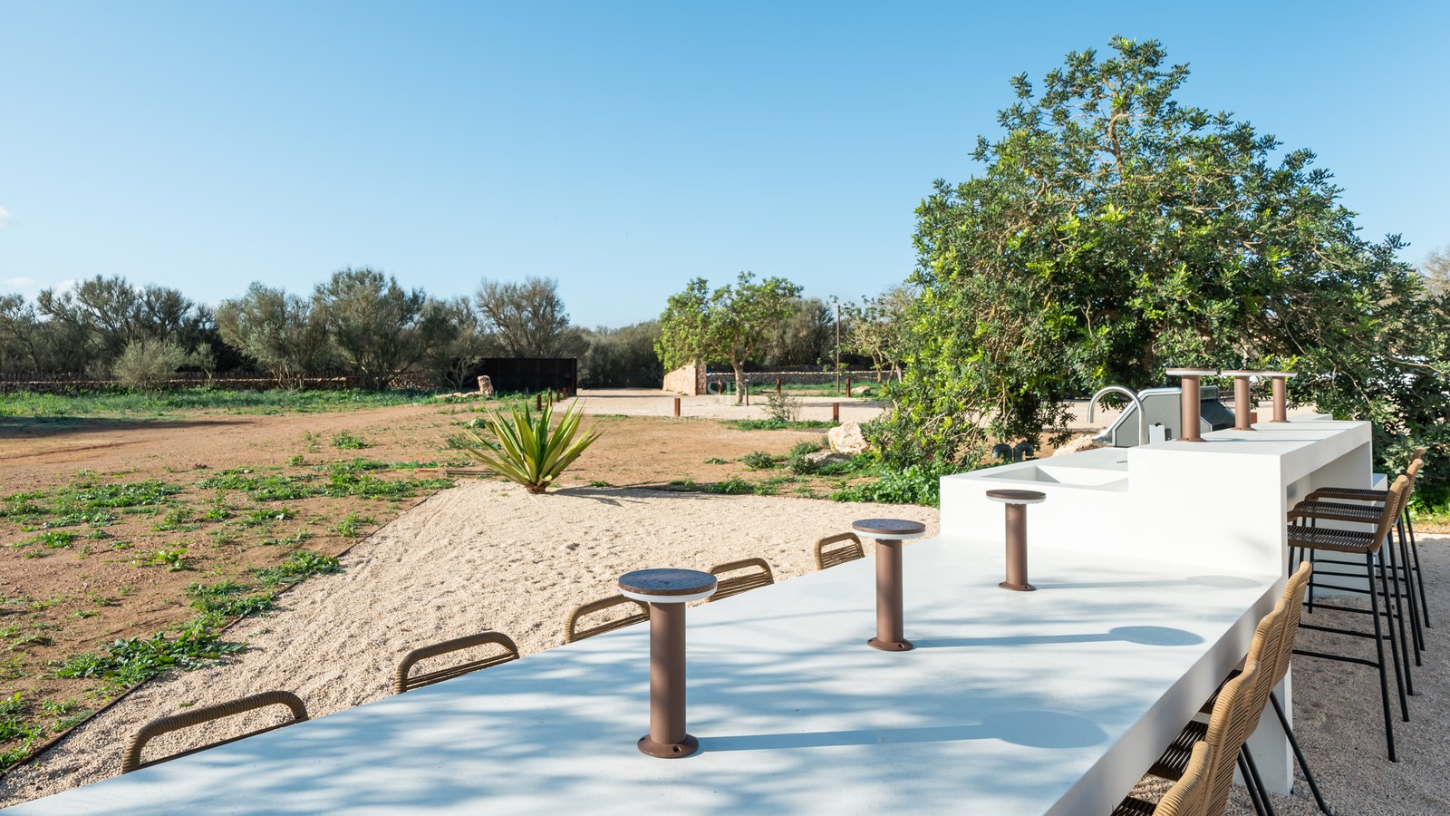 Minimalist white outdoor bar with integrated seating overlooking a Mediterranean landscape with trees and gravel.