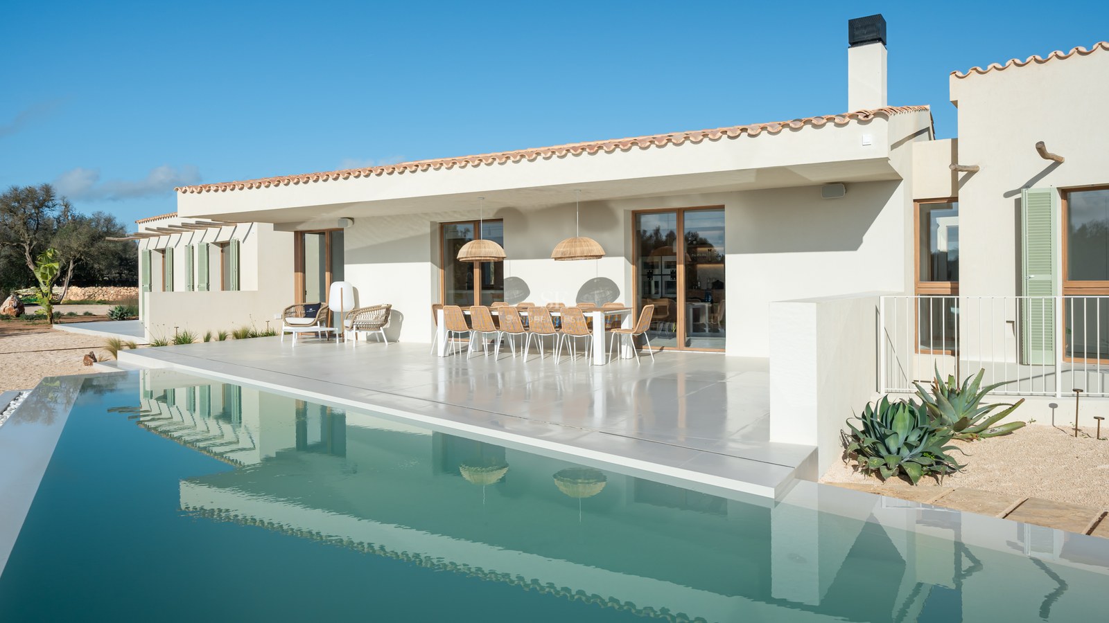 Modern white villa terrace with a large infinity pool, outdoor dining area, and clear blue sky background.