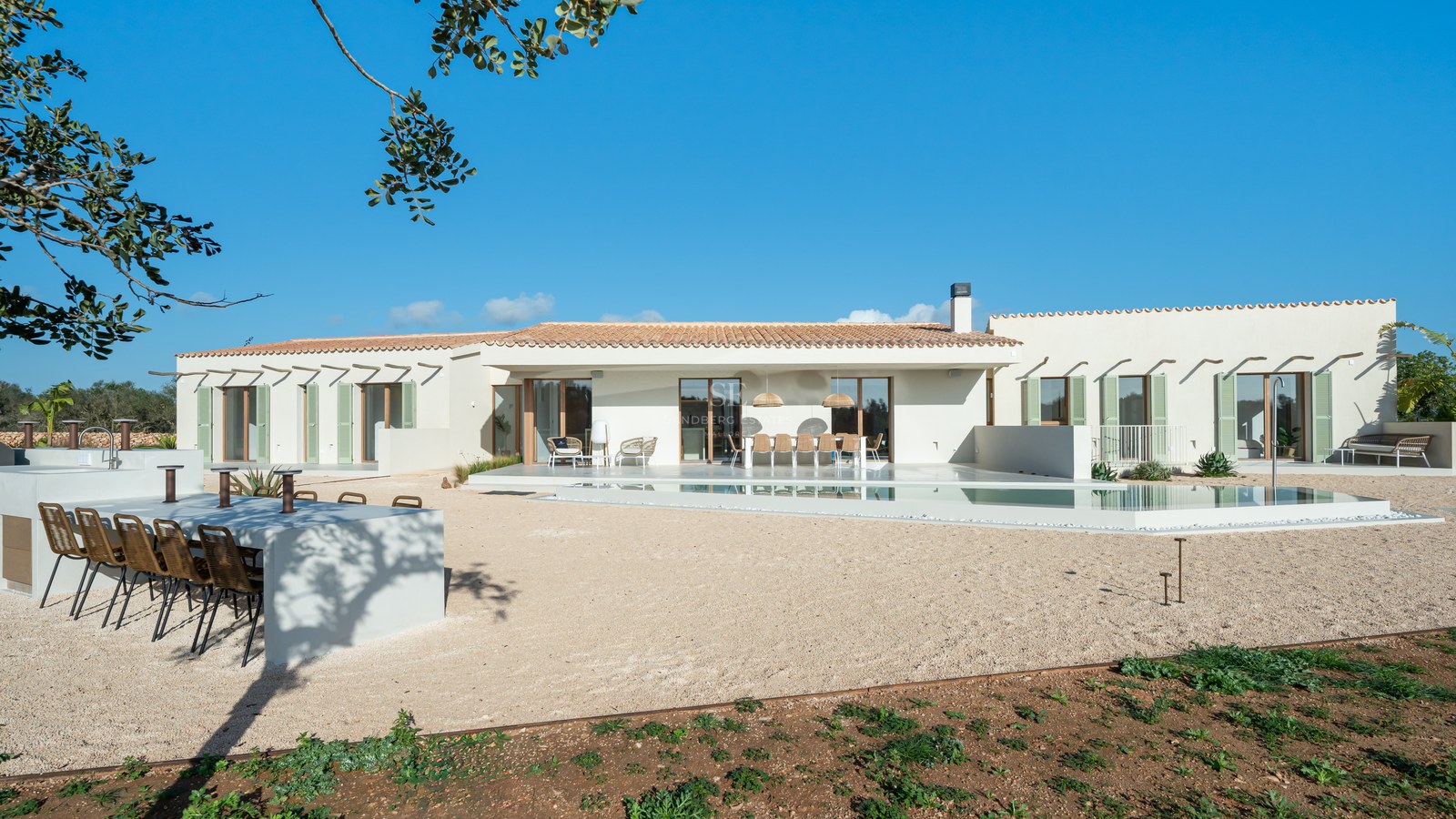 Wide shot of a beige one-story villa featuring a turquoise pool, outdoor kitchen, and green shutters under a blue sky.