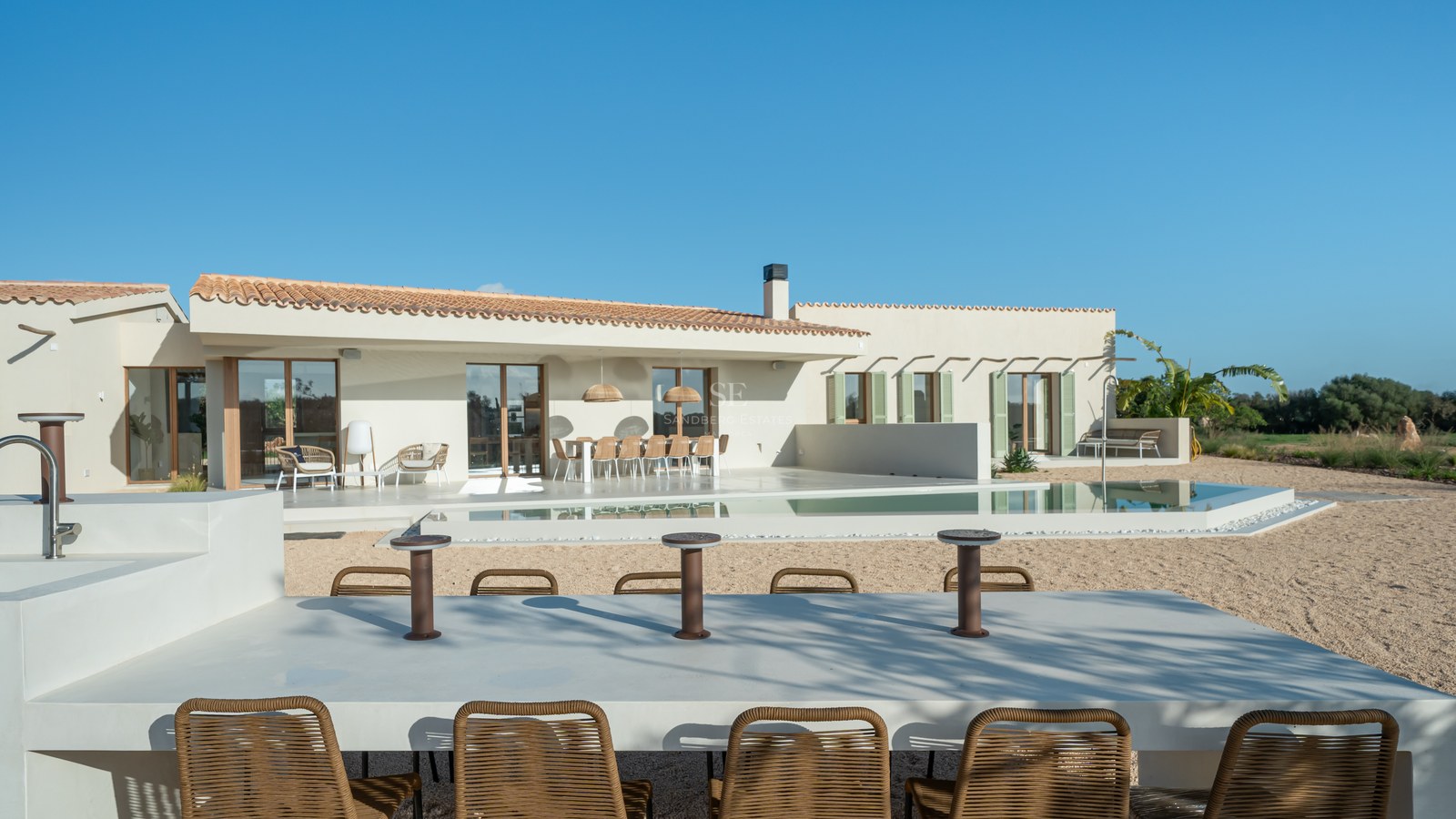 Single-story sand-colored villa with a swimming pool, multiple outdoor seating areas, and a clear blue sky.