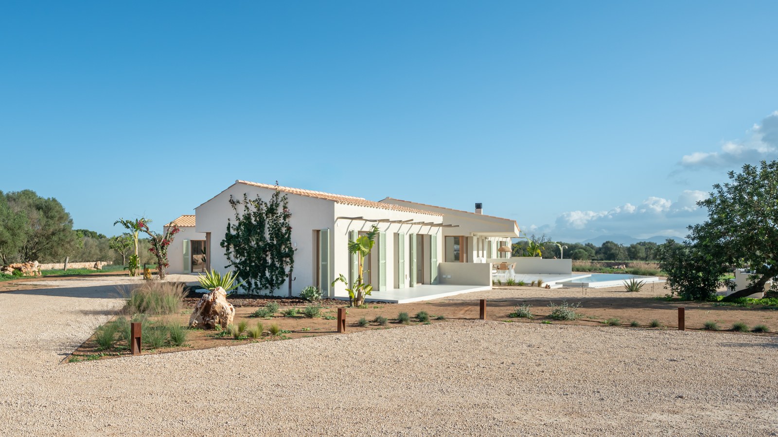 Facade of a modern villa with cream walls, sage green shutters, and a gravel driveway under a clear blue sky.