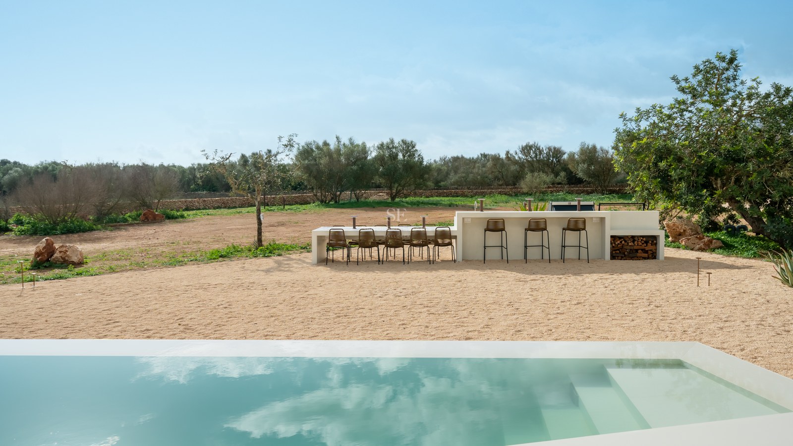 Modern white outdoor kitchen and dining area next to a sleek rectangular pool with Mediterranean landscape views.