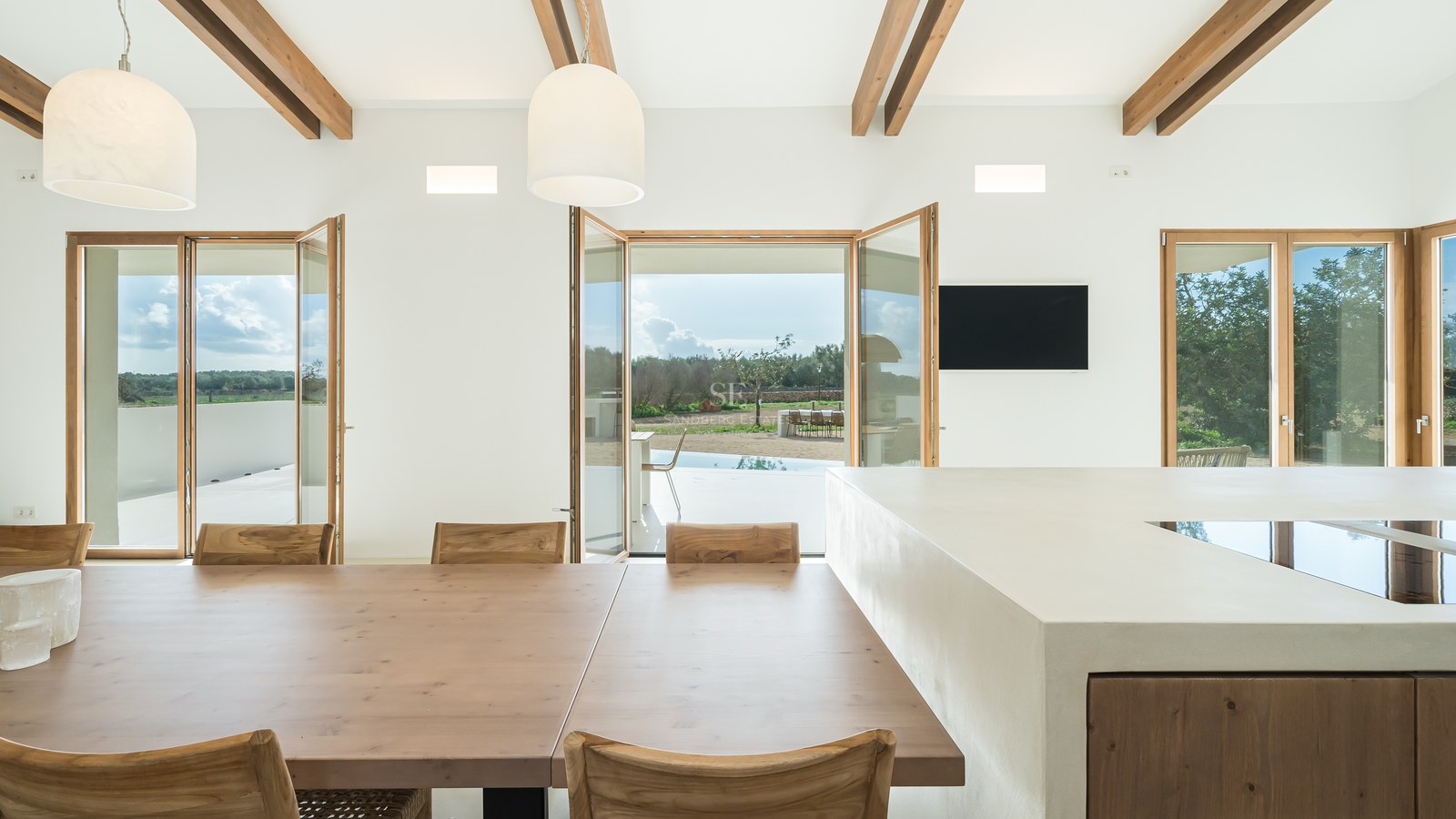 Modern kitchen with a large white island and wooden dining table featuring exposed ceiling beams and terrace views.