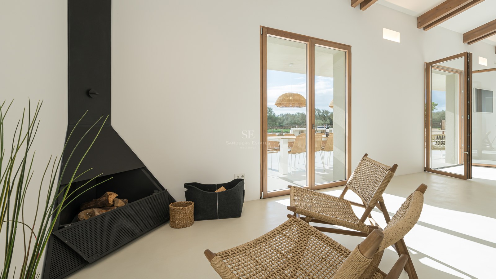 Minimalist interior featuring a sleek black fireplace, woven rattan chairs, and large wooden-framed glass doors to a terrace.