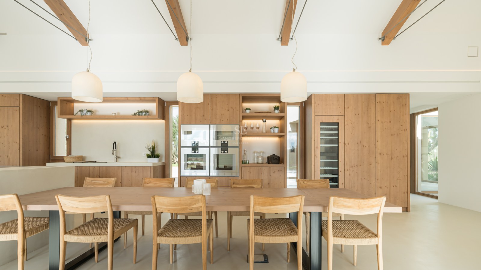 Light wood dining table with woven chairs in front of a modern oak kitchen featuring integrated appliances and wine cooler.