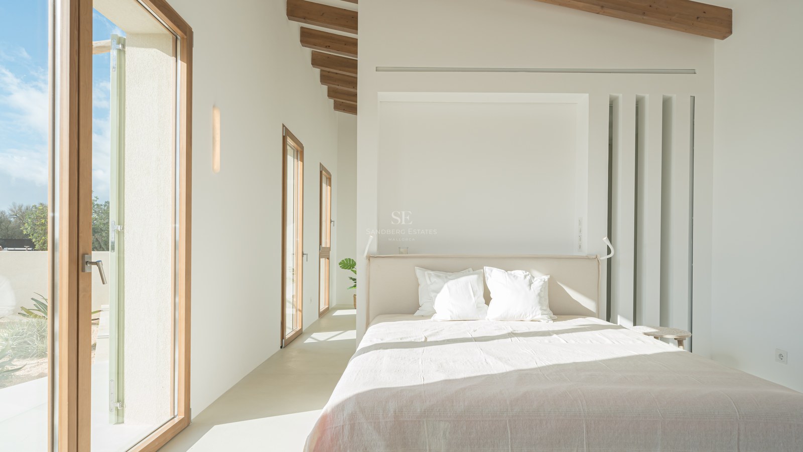 Bright master bedroom featuring exposed wooden ceiling beams, micro-cement floors, and floor-to-ceiling wooden glass doors.