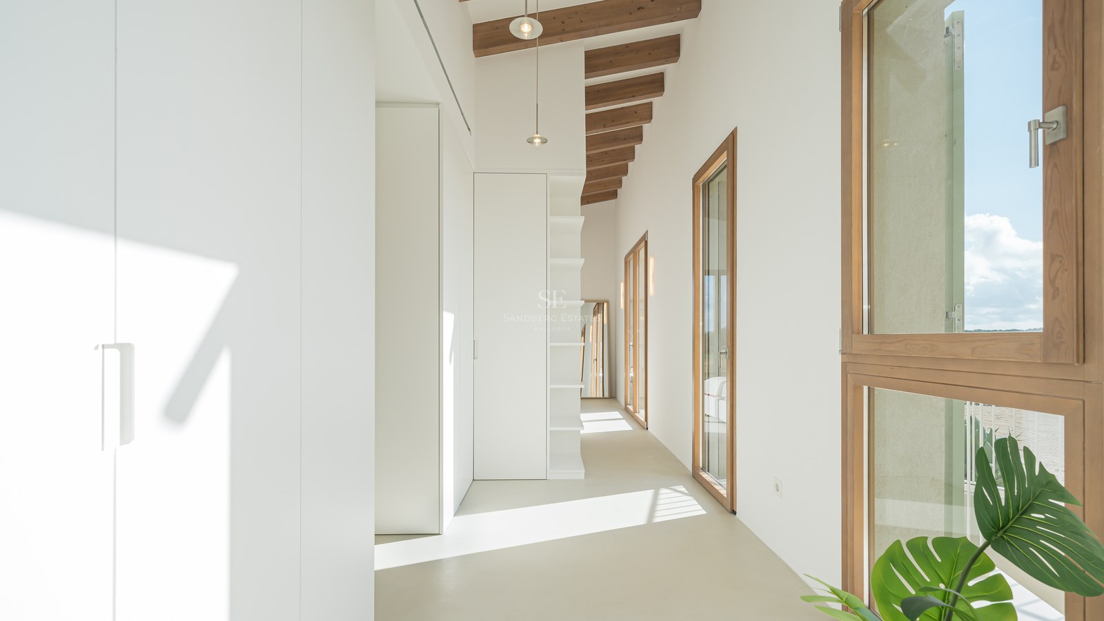 Bright white hallway featuring built-in wardrobes, exposed wooden ceiling beams, and large wood-framed windows.