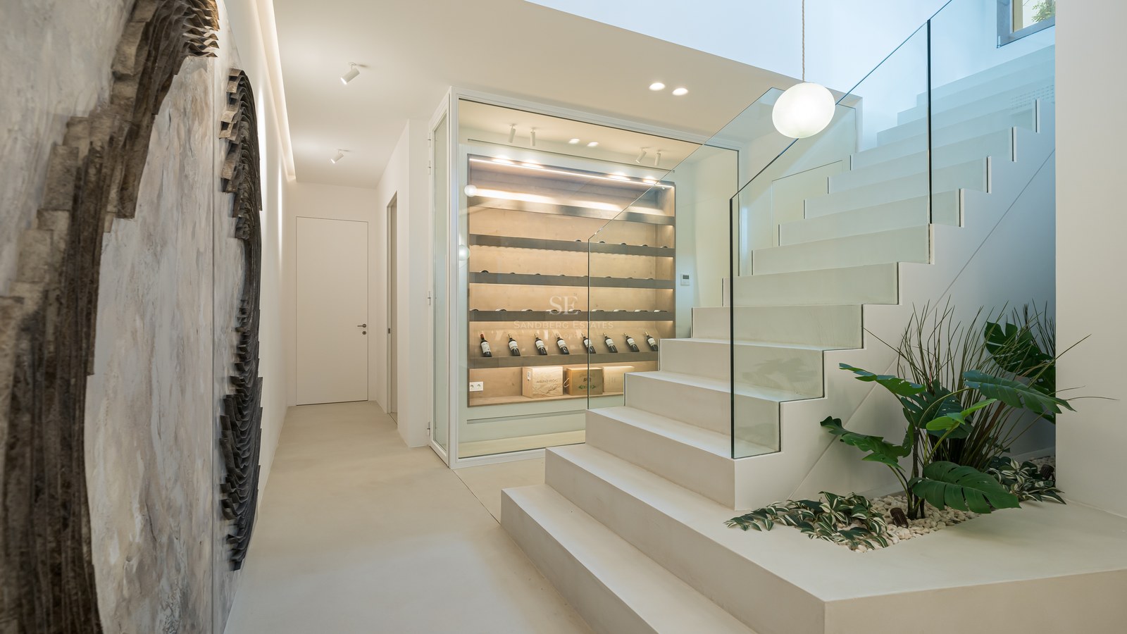 Modern white staircase with glass railing next to a glass-enclosed illuminated wine display case.