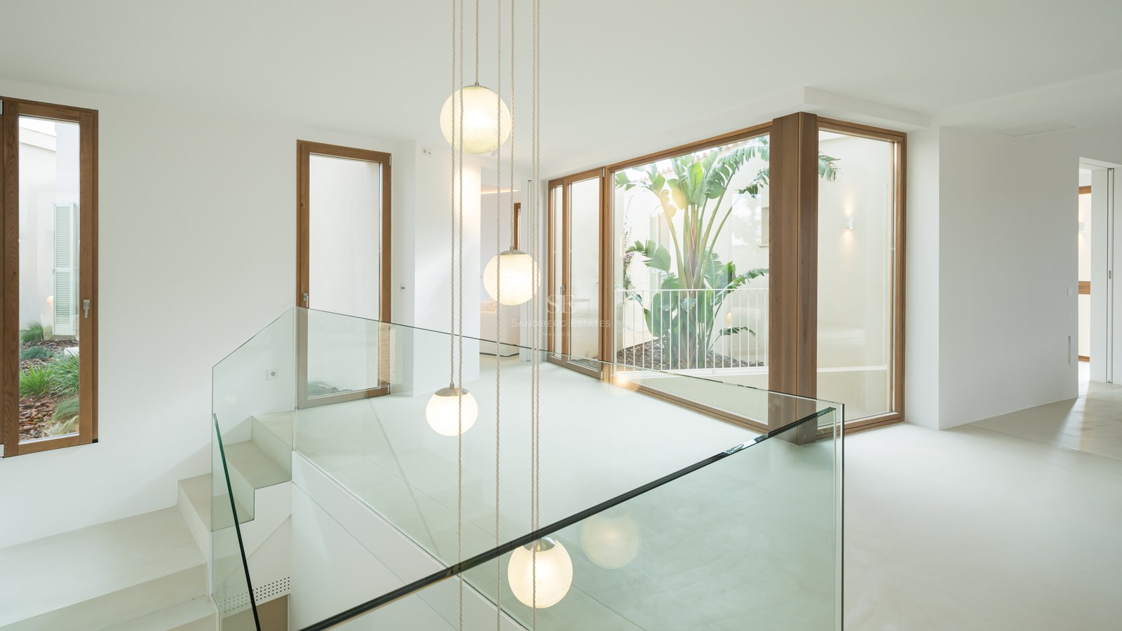 Interior view of a modern hallway with glass railings, wooden window frames, and hanging globe lights over a patio.