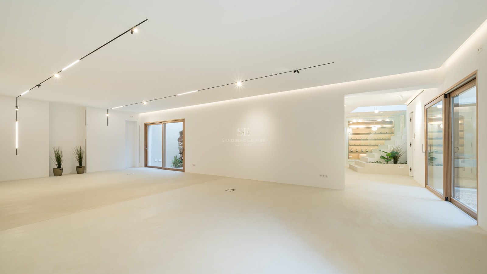 Large empty open-plan room with white walls, polished cream floors, track lighting, and wood-framed glass doors.