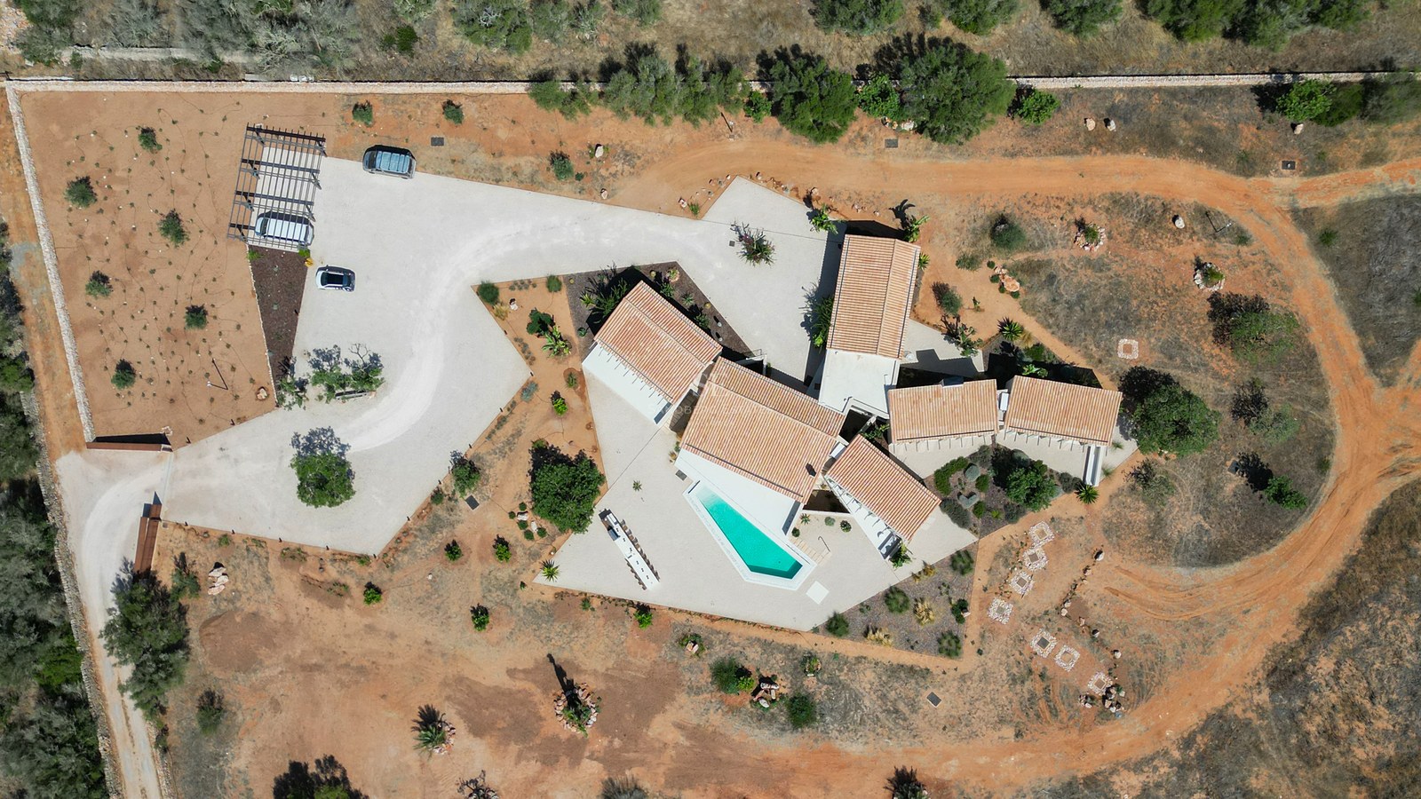 High-angle drone shot of a white modern villa with terracotta roofs, a geometric pool, and red soil grounds.