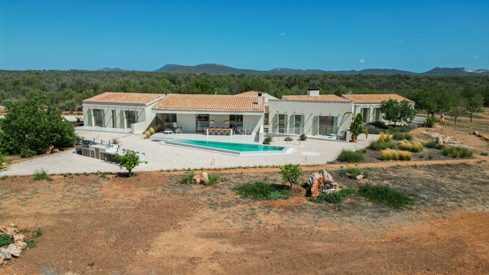 Elevated view of a modern single-story villa with a turquoise pool, outdoor kitchen, and terracotta roof in Mallorca.