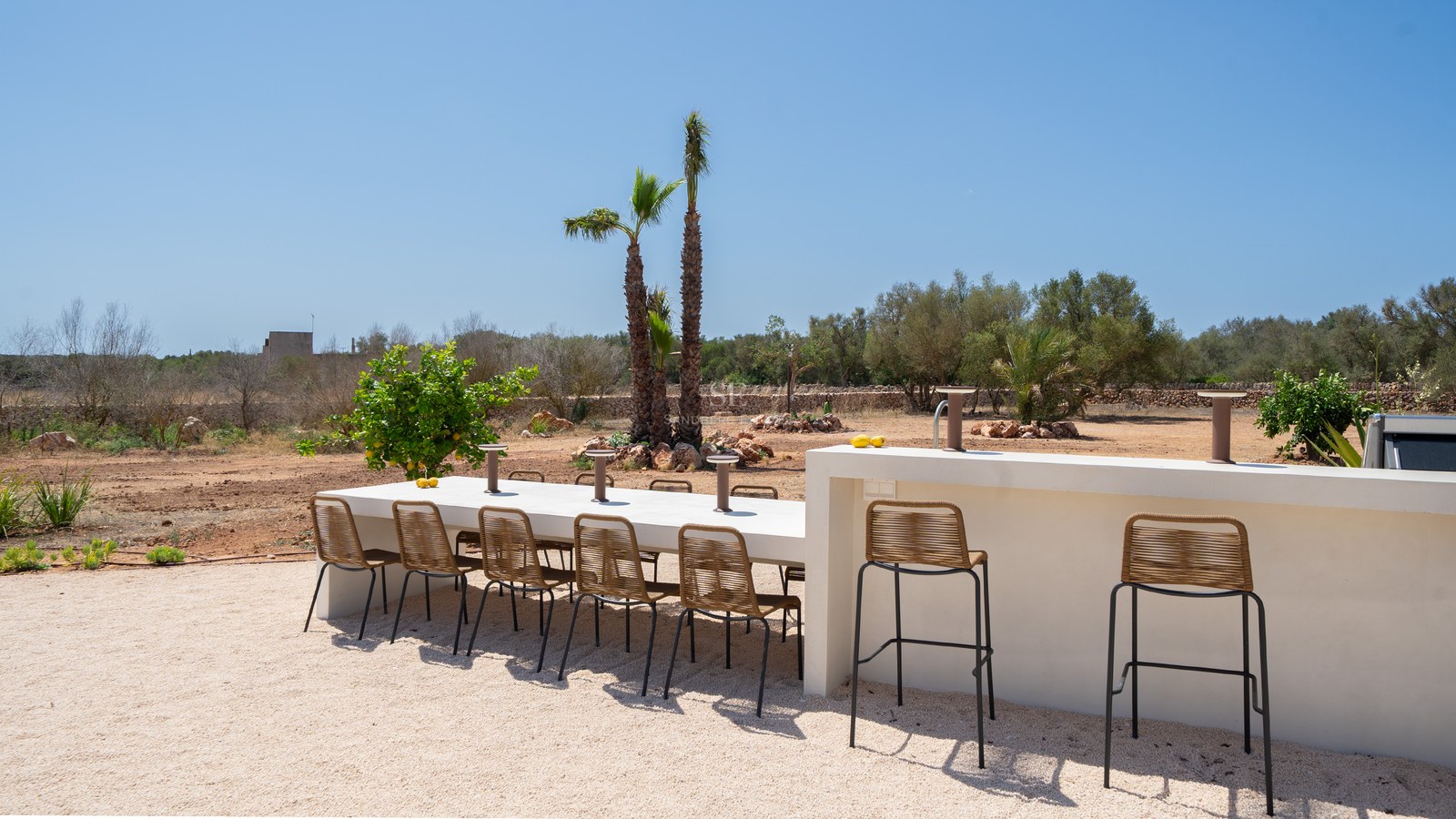 A long white dining table and bar counter with woven chairs on a gravel terrace under a clear blue sky.