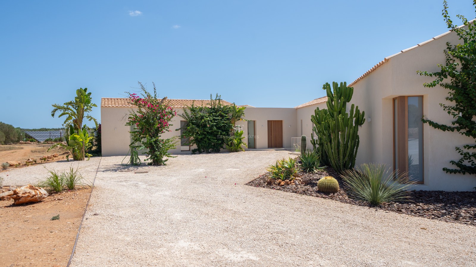 Contemporary cream-colored villa exterior with gravel courtyard, wooden doors, and desert-style landscaping under a clear sky.
