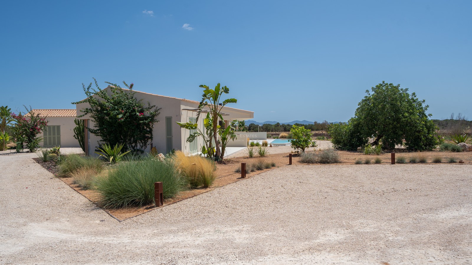 A modern one-story white villa with a terracotta roof, gravel driveway, and lush Mediterranean plants under a clear sky.