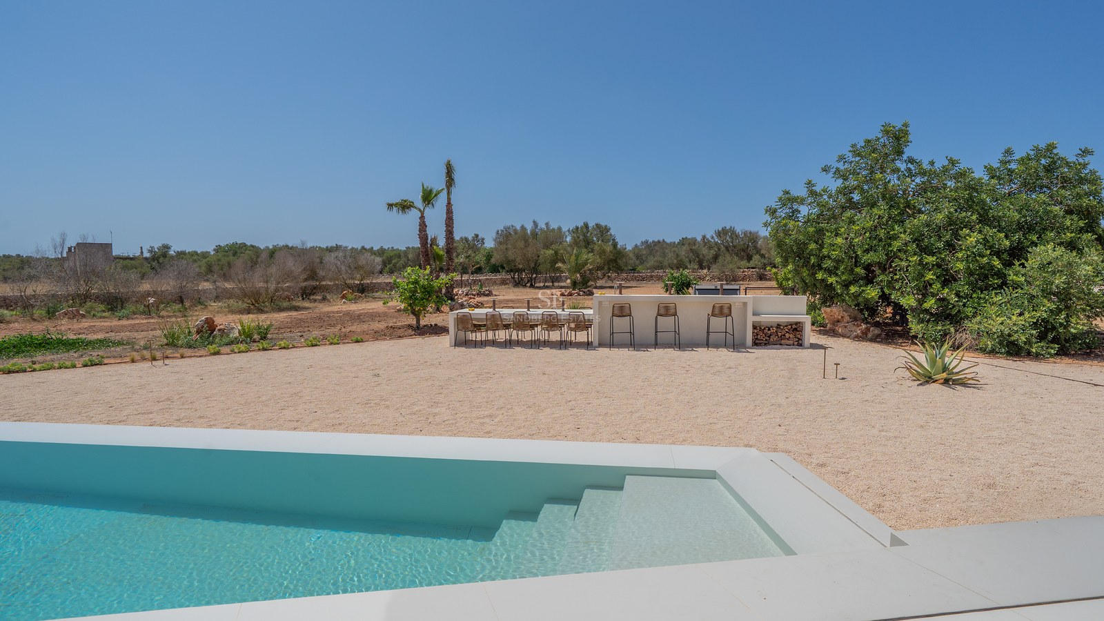 Modern white swimming pool foreground with outdoor dining area and bar set against a Mediterranean landscape.