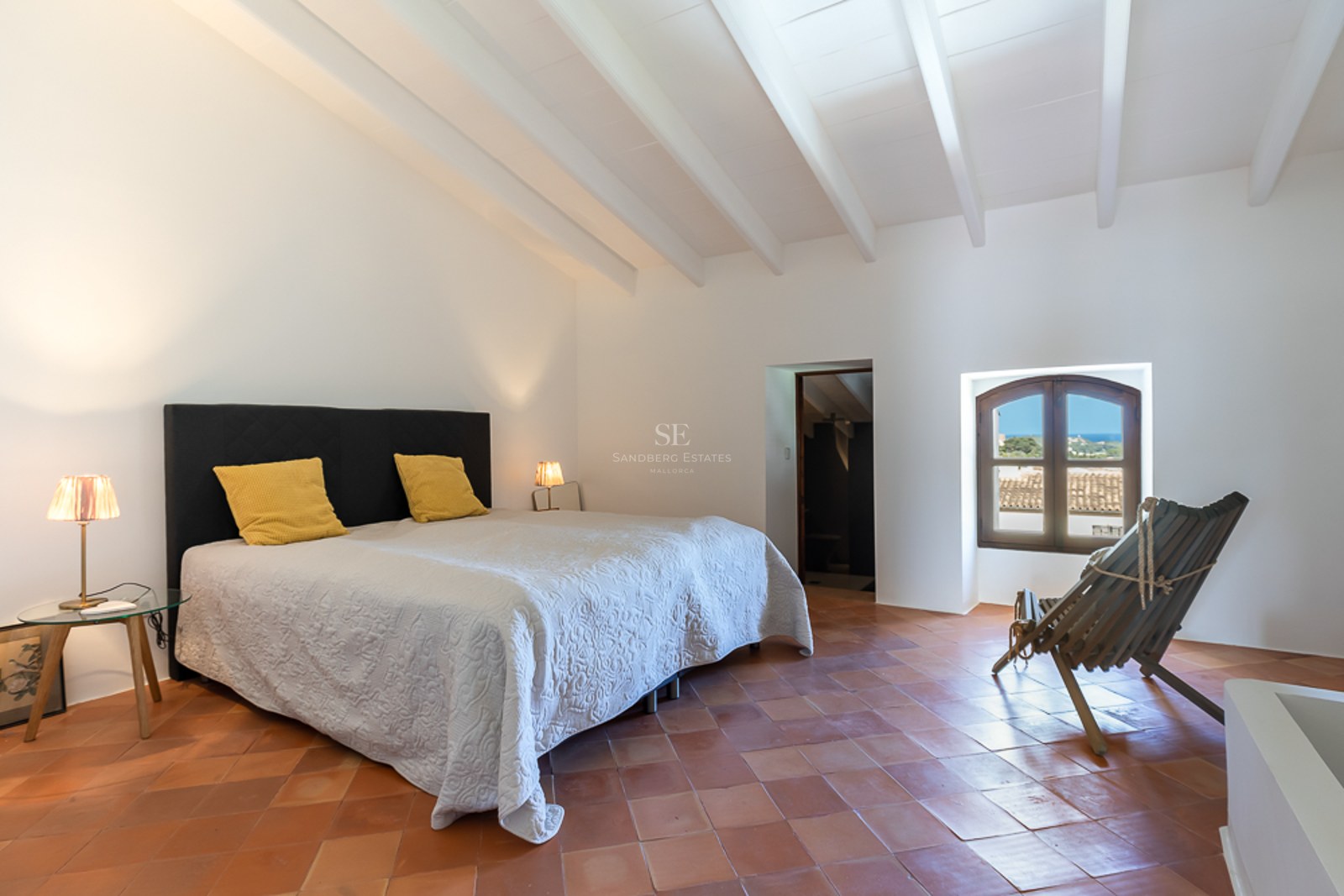 Schlafzimmer mit weißer Balkendecke, Terrakottaboden, großem Bett und Rundbogenfenster mit Blick auf das Meer.