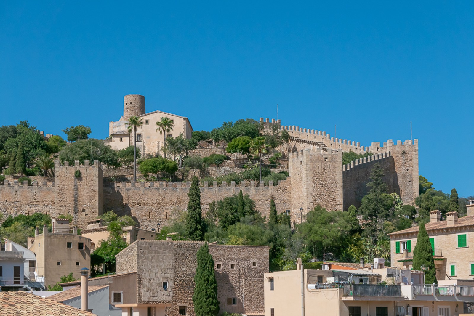 Steinmauern und Türme einer mittelalterlichen Festung auf einem Hügel unter blauem Himmel in einem mallorquinischen Dorf.
