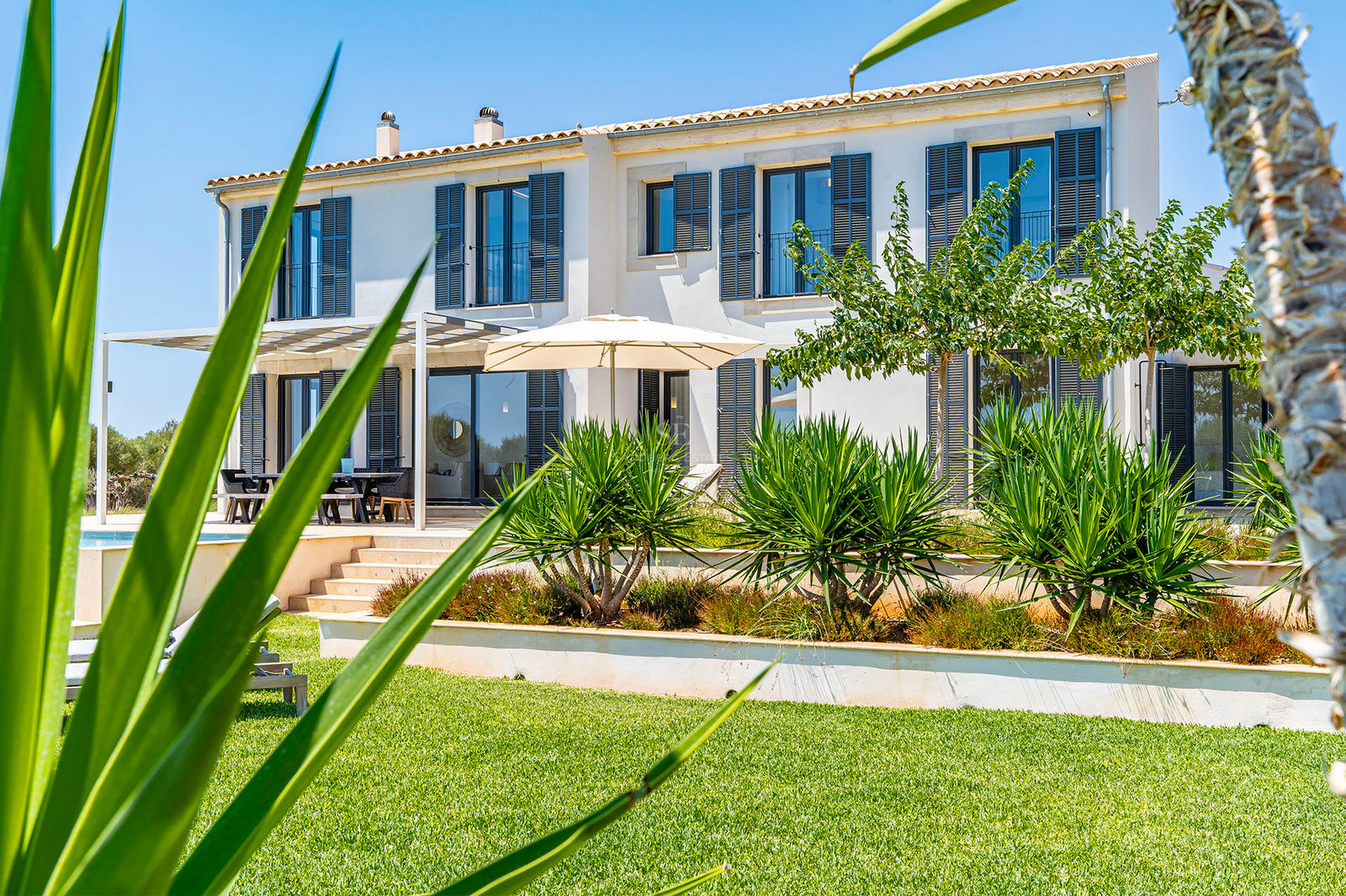 Villa blanche à deux étages avec volets gris, jardins en terrasses et pelouse soignée sous un ciel bleu.