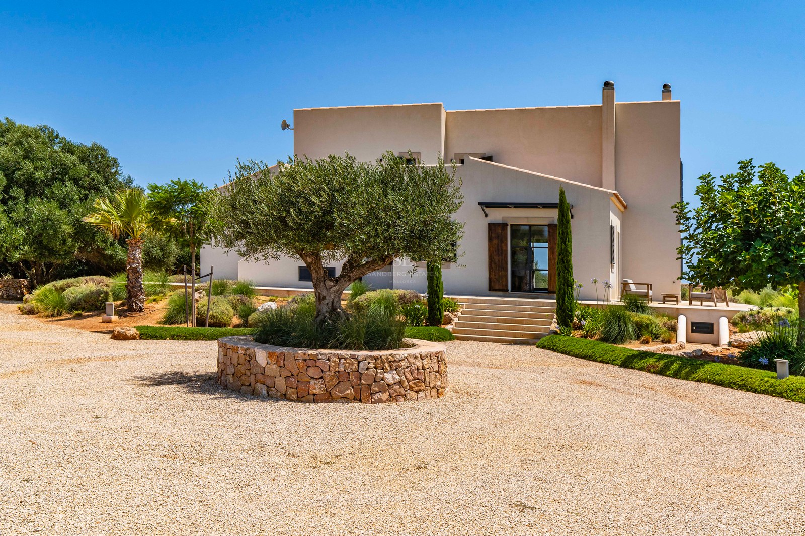 Entrée de villa moderne beige avec un grand olivier dans un muret en pierre et une allée de gravier.