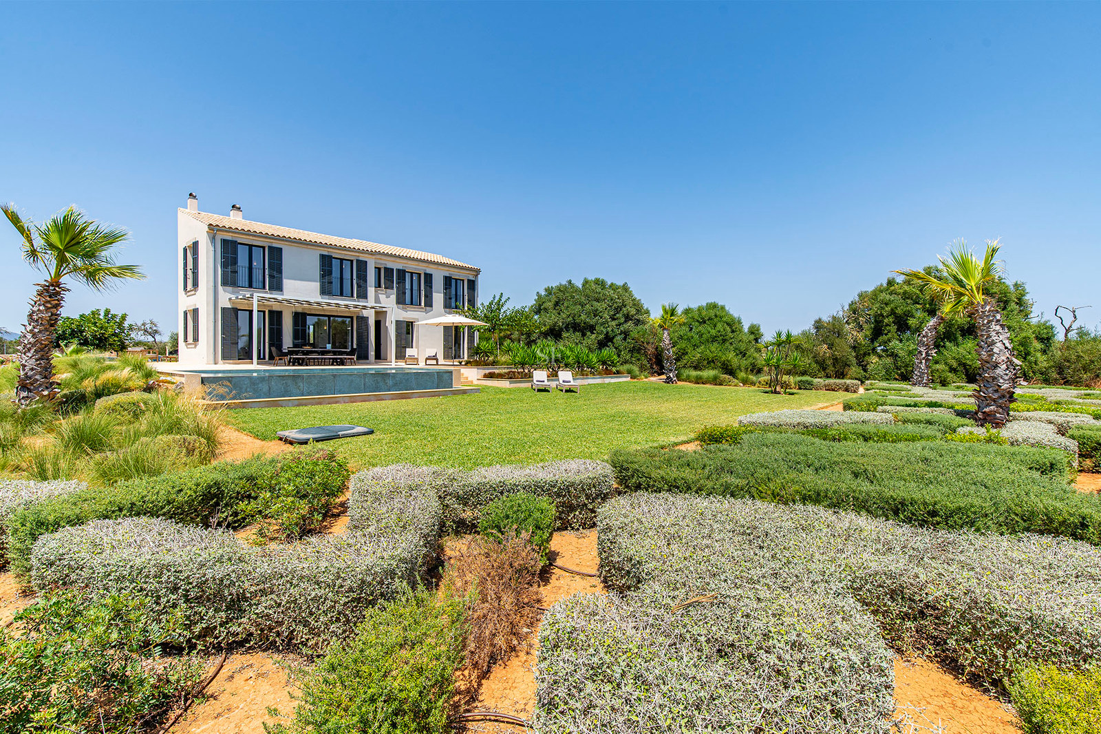 Villa de deux étages aux volets sombres, piscine et jardins soignés sous un ciel bleu dégagé.