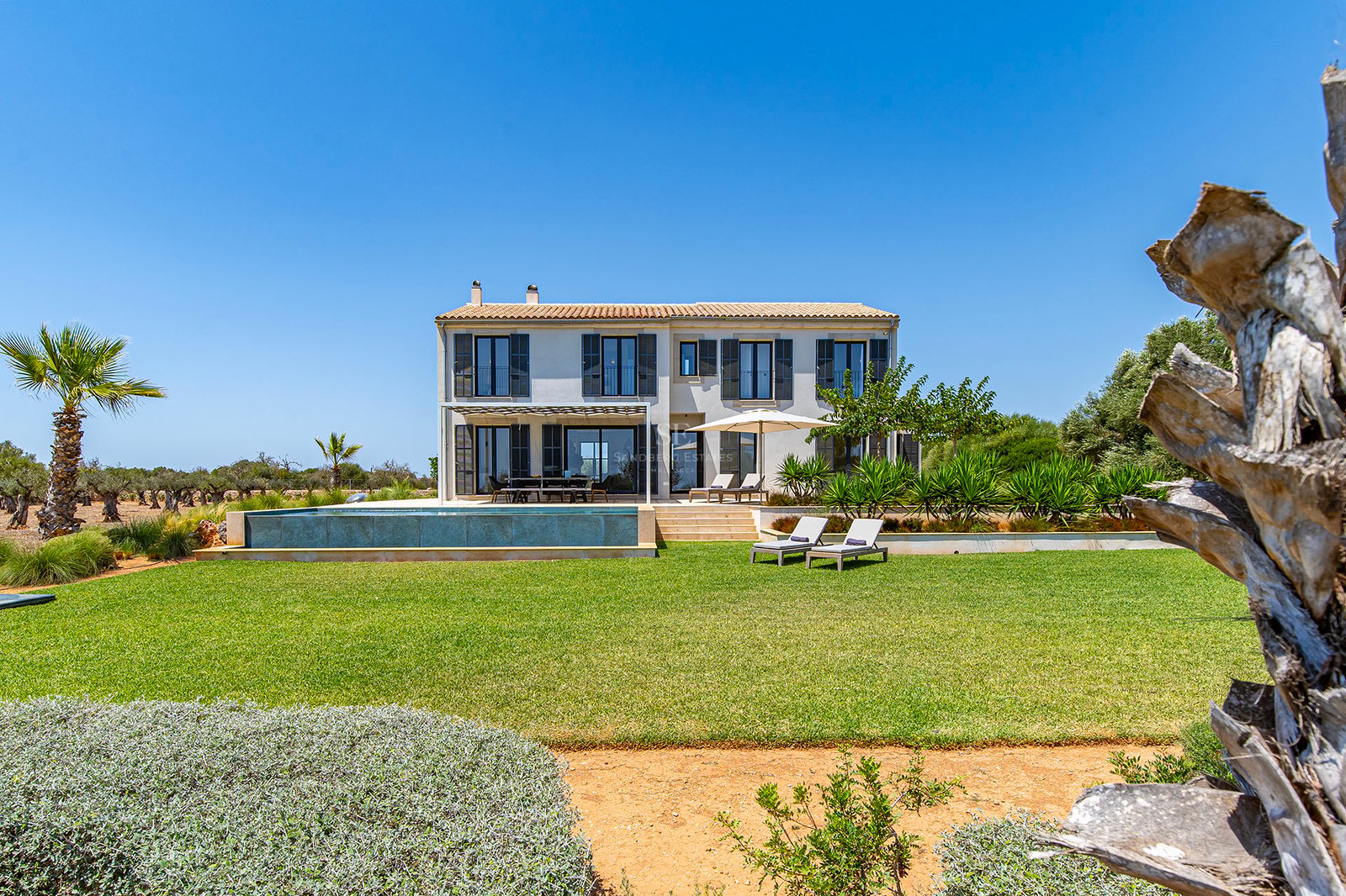 Villa beige à deux étages avec piscine à débordement, pelouse verte et volets sombres par temps clair.
