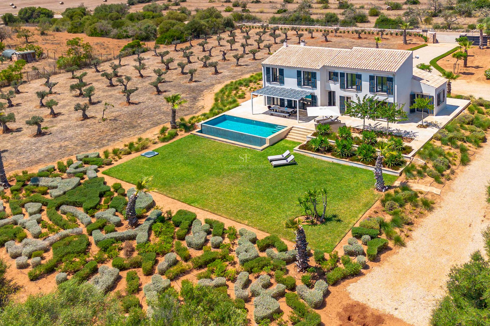 Vue aérienne d'une villa blanche à deux étages avec piscine à débordement, pelouse soignée et oliveraies environnantes.