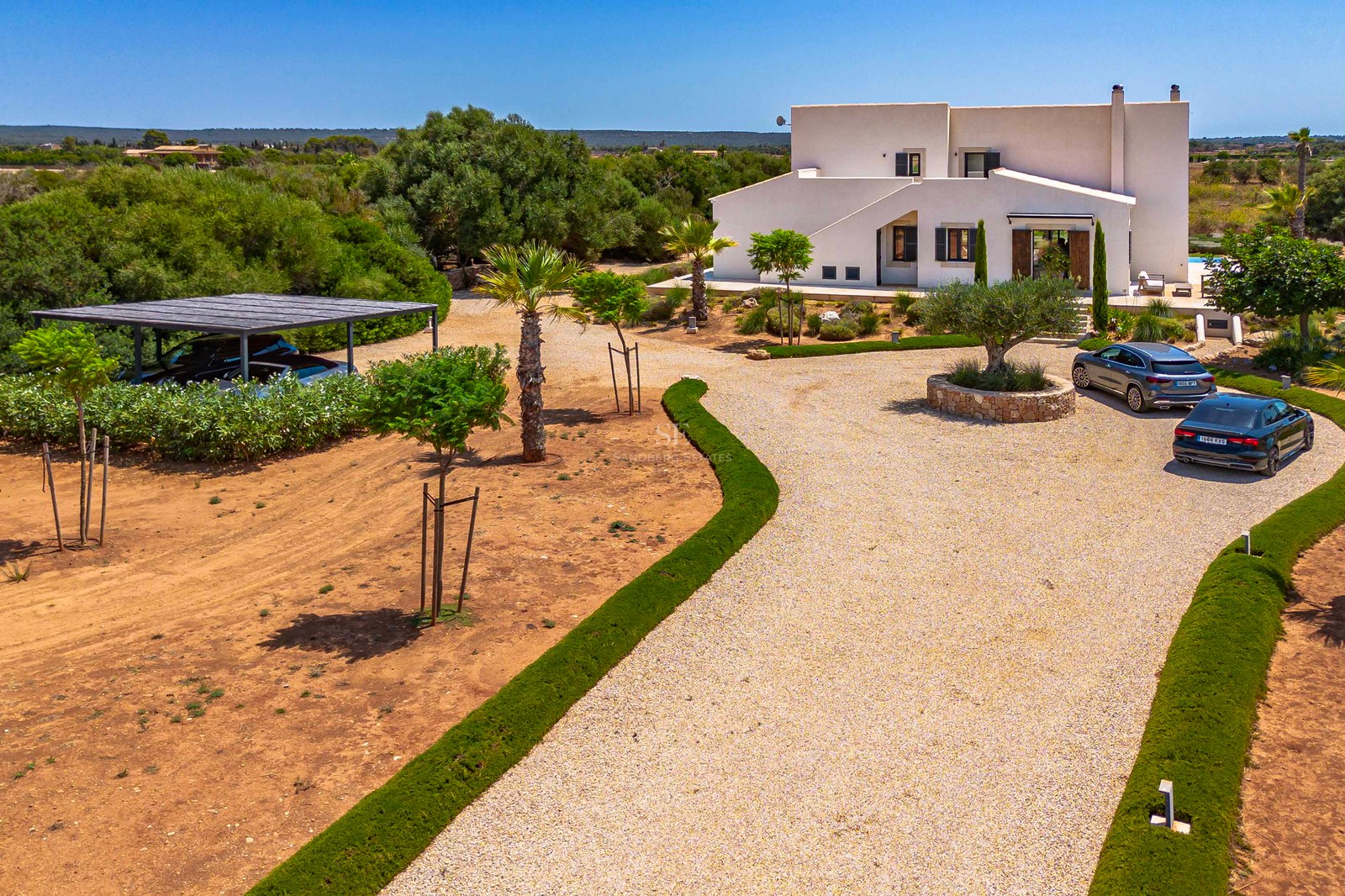 Vue aérienne d'une villa blanche moderne avec une allée en gravier, des jardins et un abri pour voitures.