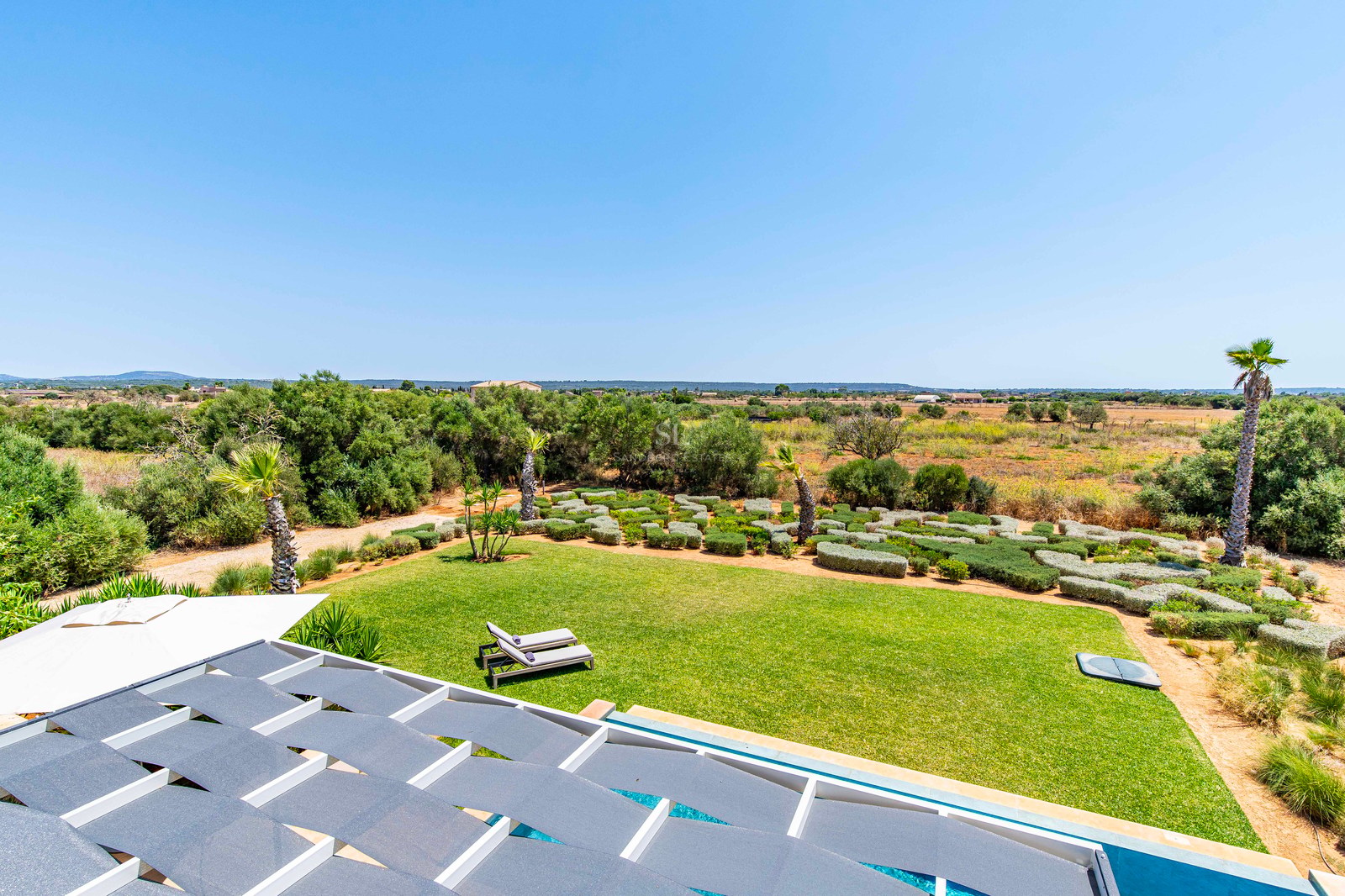 Vue depuis une terrasse avec pergola sur un vaste jardin paysager, des palmiers et la campagne environnante.