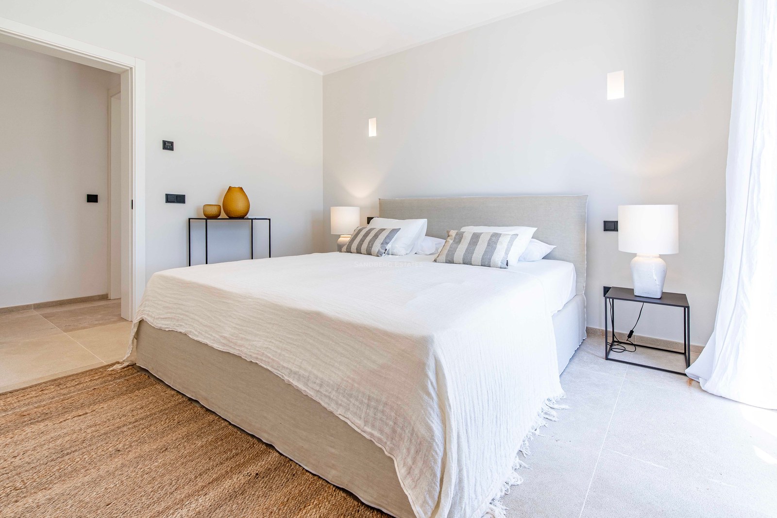 Chambre minimaliste avec sol en pierre, tapis en jute, grand lit et lampes de chevet modernes.