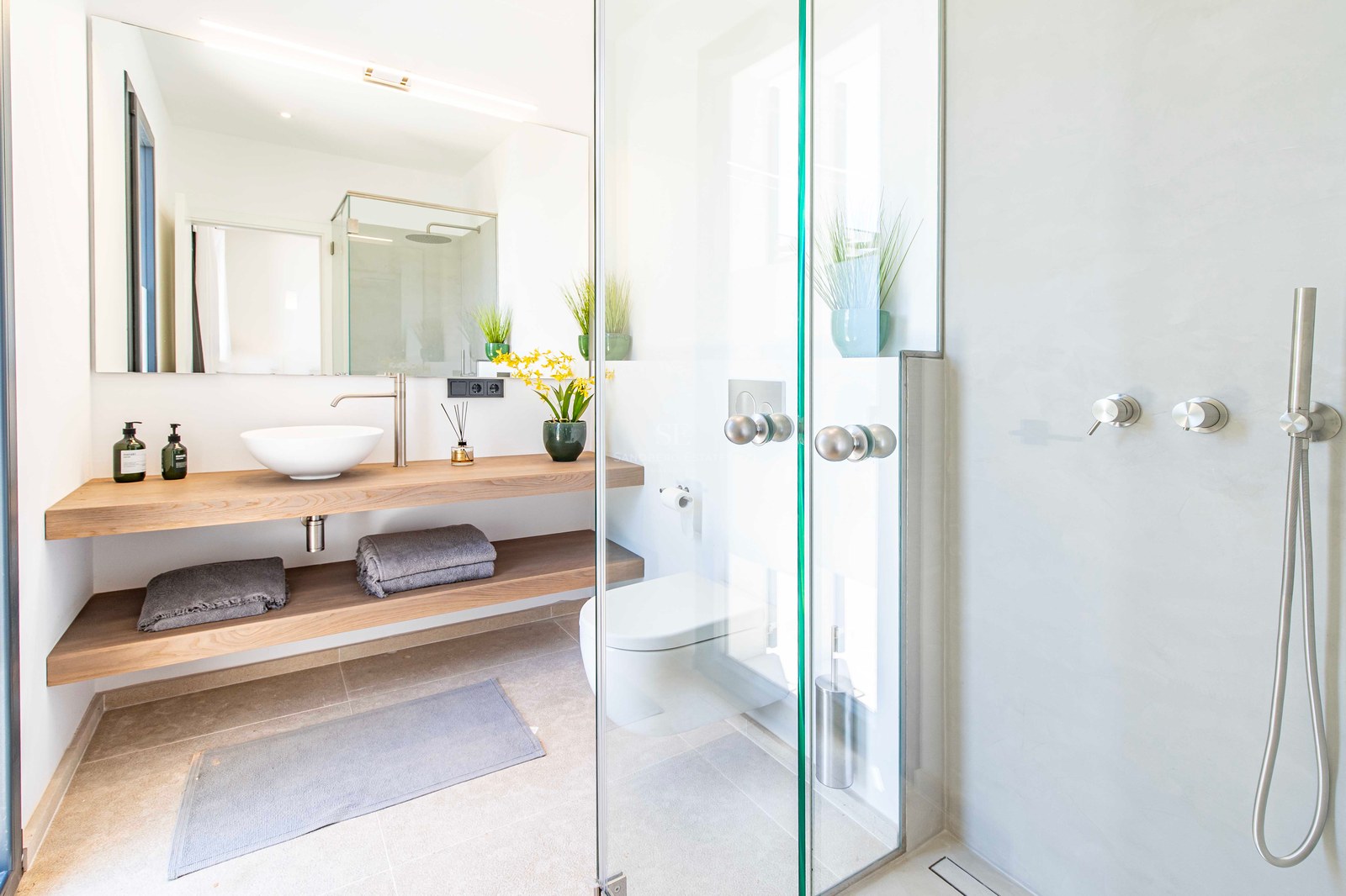 Salle de bains lumineuse avec douche à l'italienne, étagères en chêne, vasque blanche et toilettes suspendues.