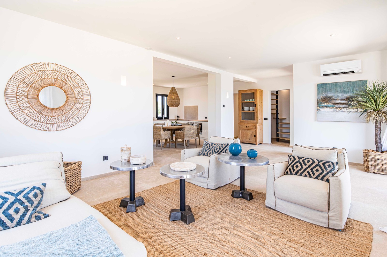 Salon lumineux avec murs blancs, sol en pierre, tapis en jute, fauteuils beiges et tables d'appoint en métal.
