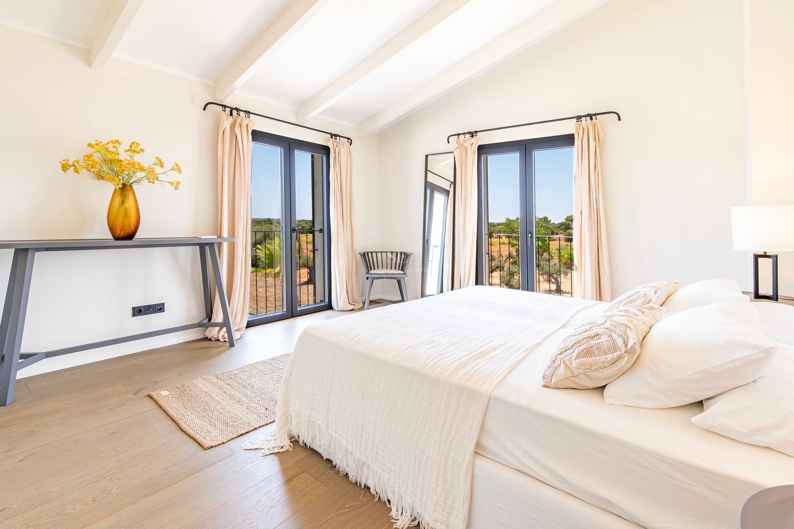 Chambre parentale lumineuse avec plafond voûté à poutres blanches, deux portes-fenêtres sombres et parquet en chêne.