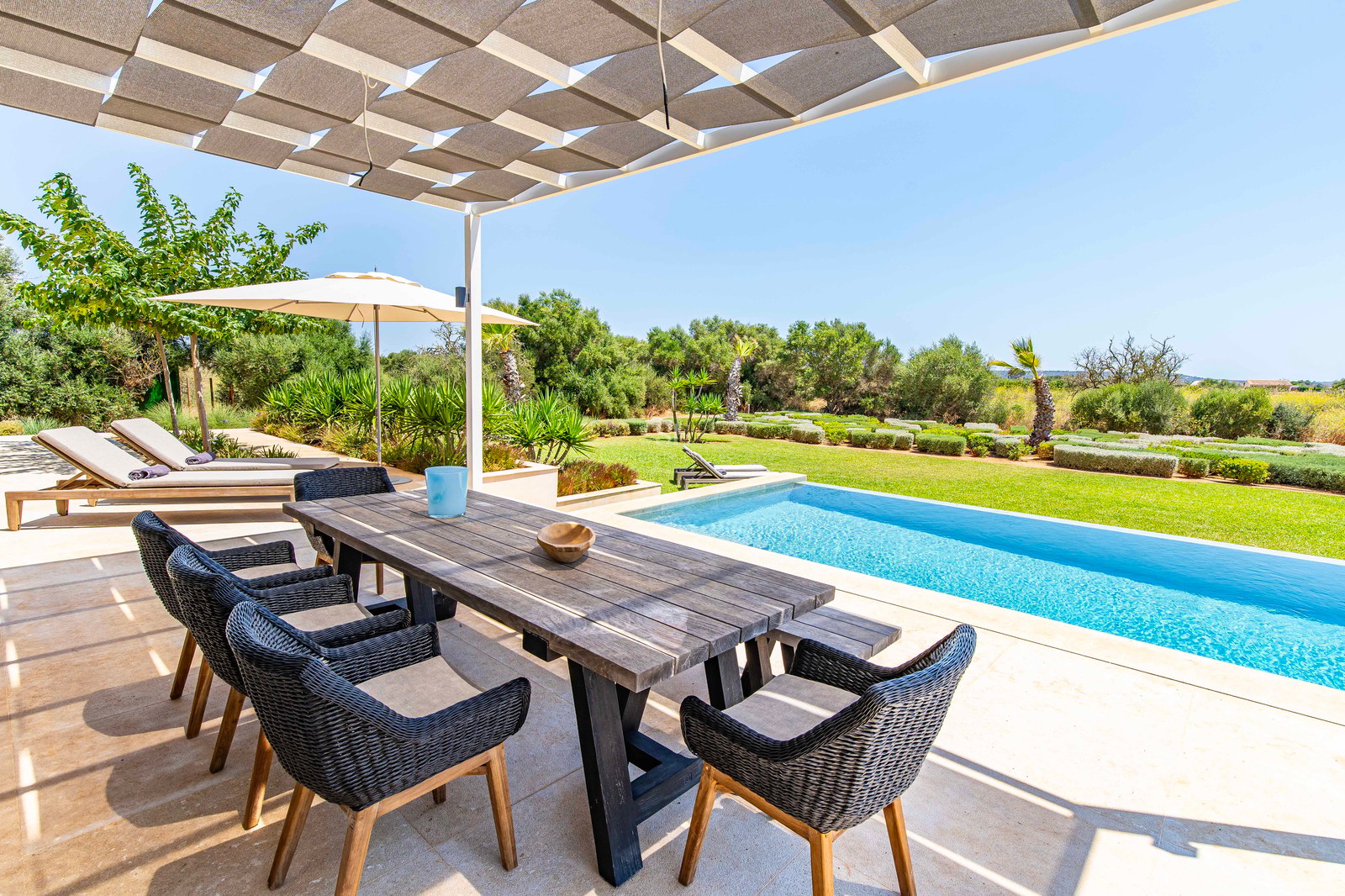 Espace repas extérieur avec table en bois et chaises en osier sous un voile d'ombrage, face à une piscine et un jardin.