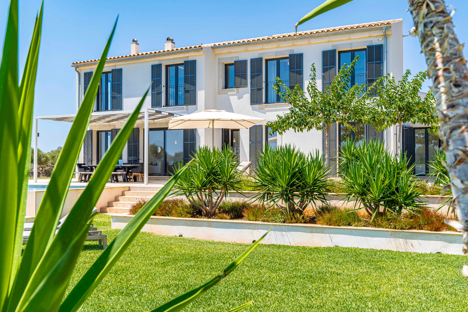 Villa blanche à deux étages avec volets bleus et jardins en terrasses sous un ciel bleu azur.