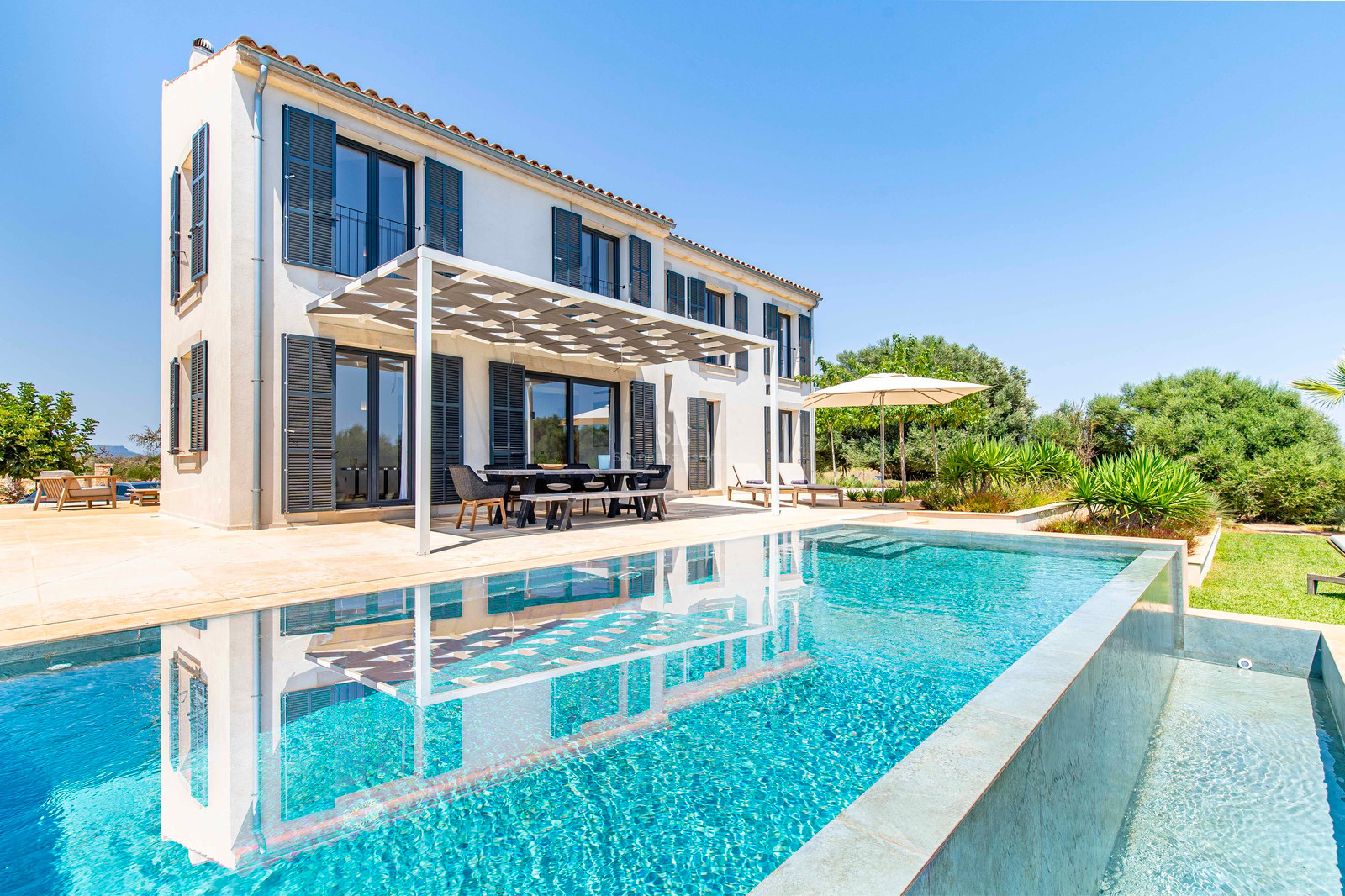 Extérieur d'une villa de luxe avec piscine turquoise et terrasse ombragée sous un ciel bleu azur.