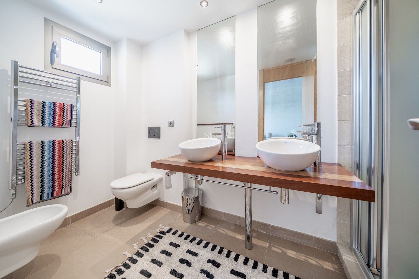 Bright bathroom with a wooden floating vanity, two white vessel sinks, heated towel rail, and large mirrors.
