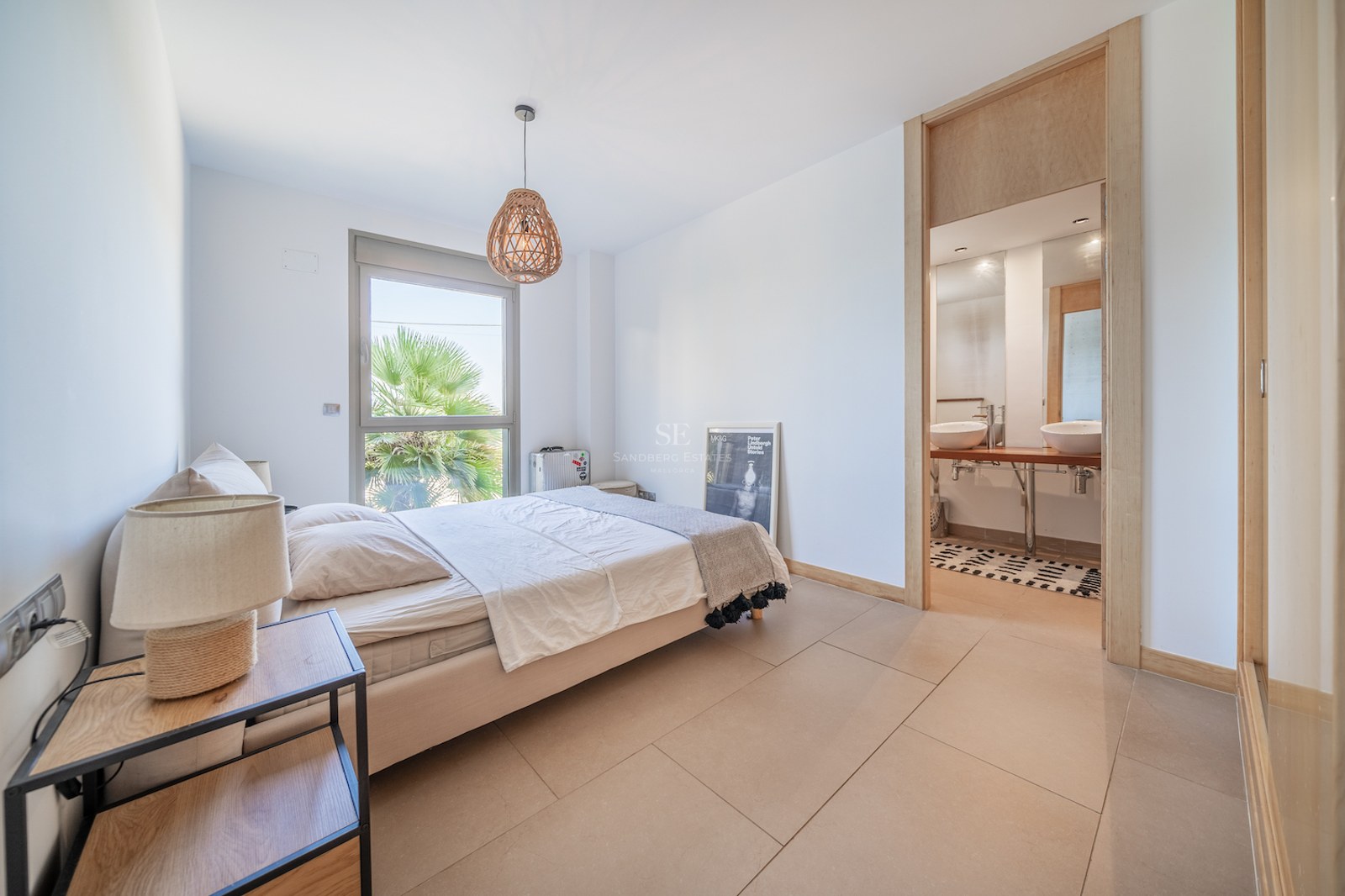 Bright modern bedroom with light wood accents, an ensuite bathroom with double sinks, and a window showing palm trees.
