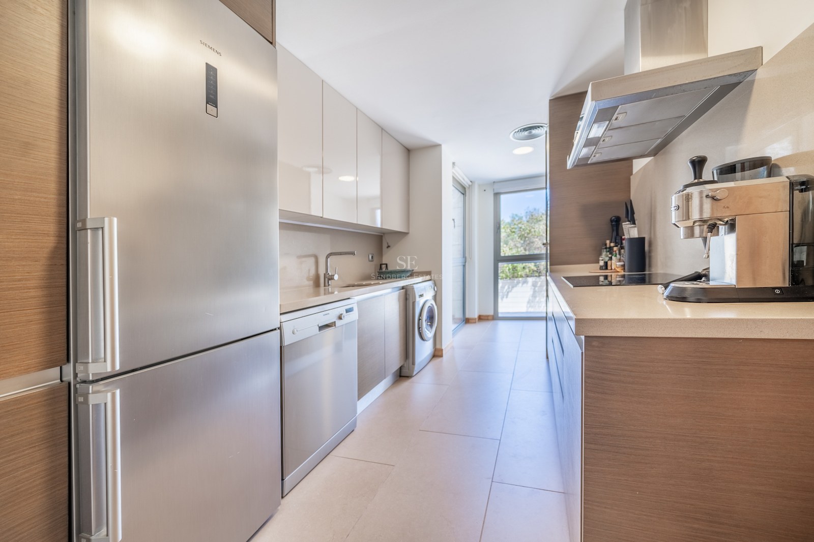 Galley-style kitchen with light wood cabinetry, stainless steel appliances, and large window at the end.