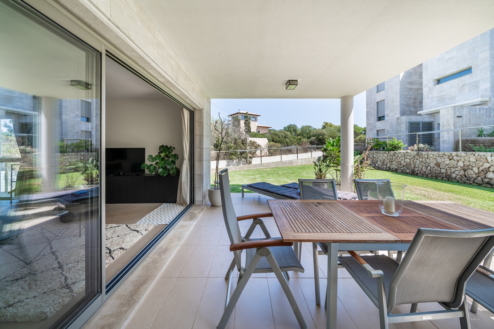 Covered terrace with wooden dining table and chairs leading to a private lawn and natural stone walls.