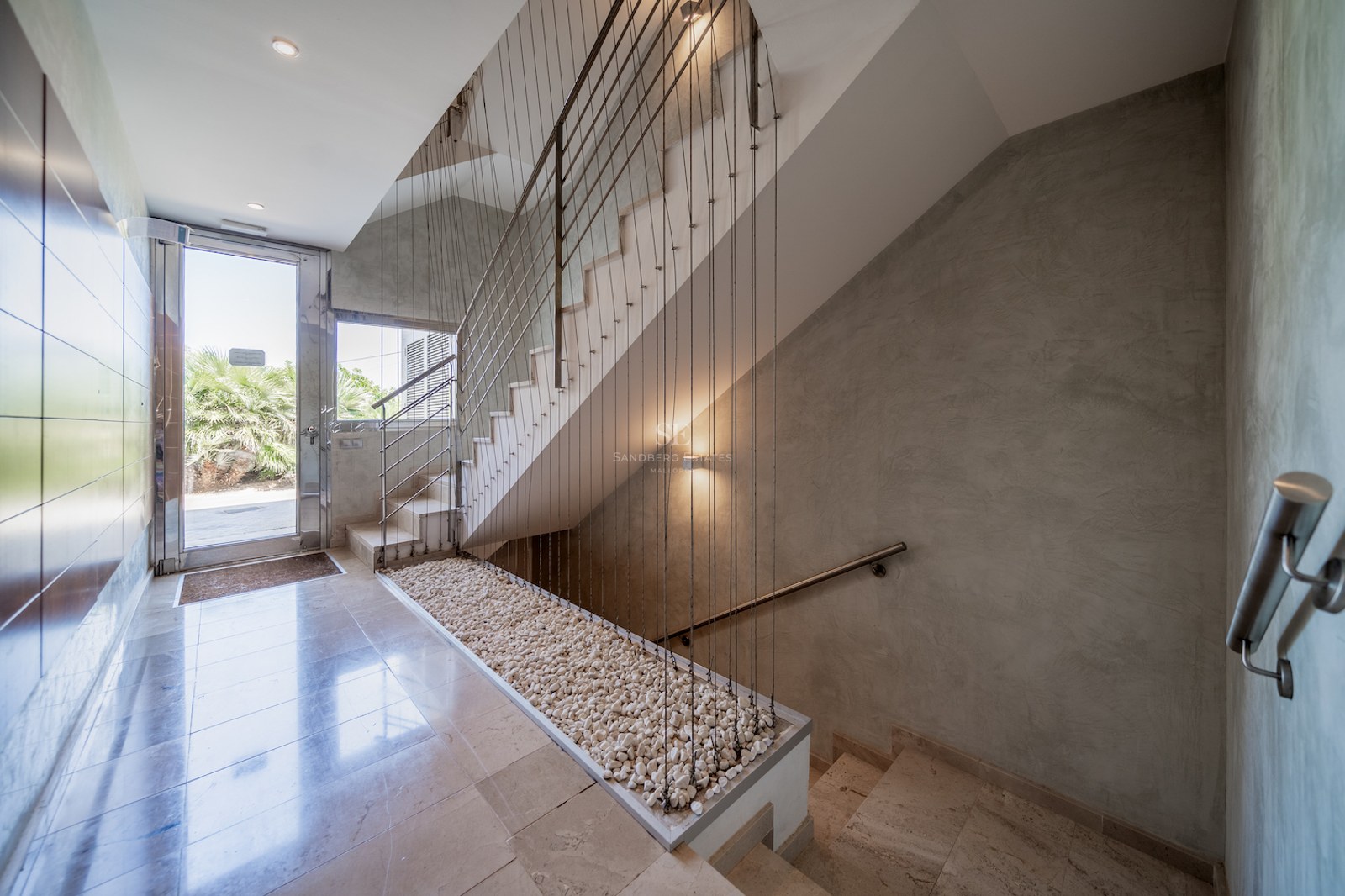 Bright entrance featuring polished stone floors, a staircase with cable railings, and a glass door to a garden.