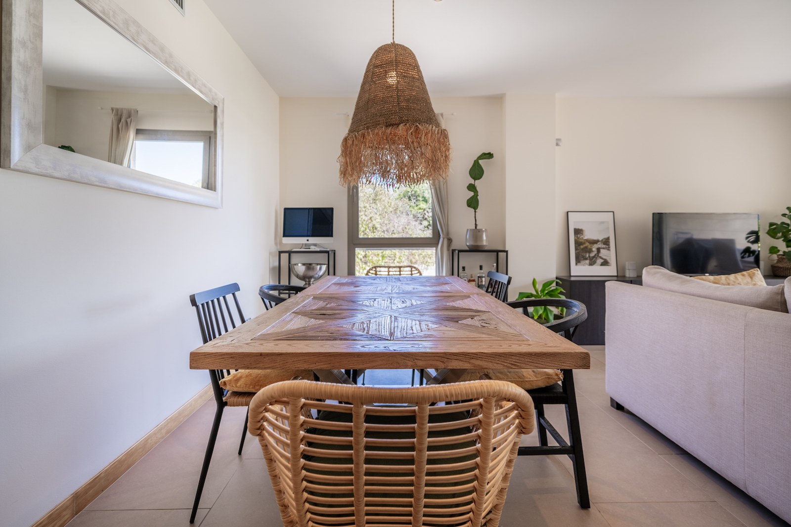 A large wooden parquetry dining table with a rattan pendant light in a bright, modern open-plan living room.