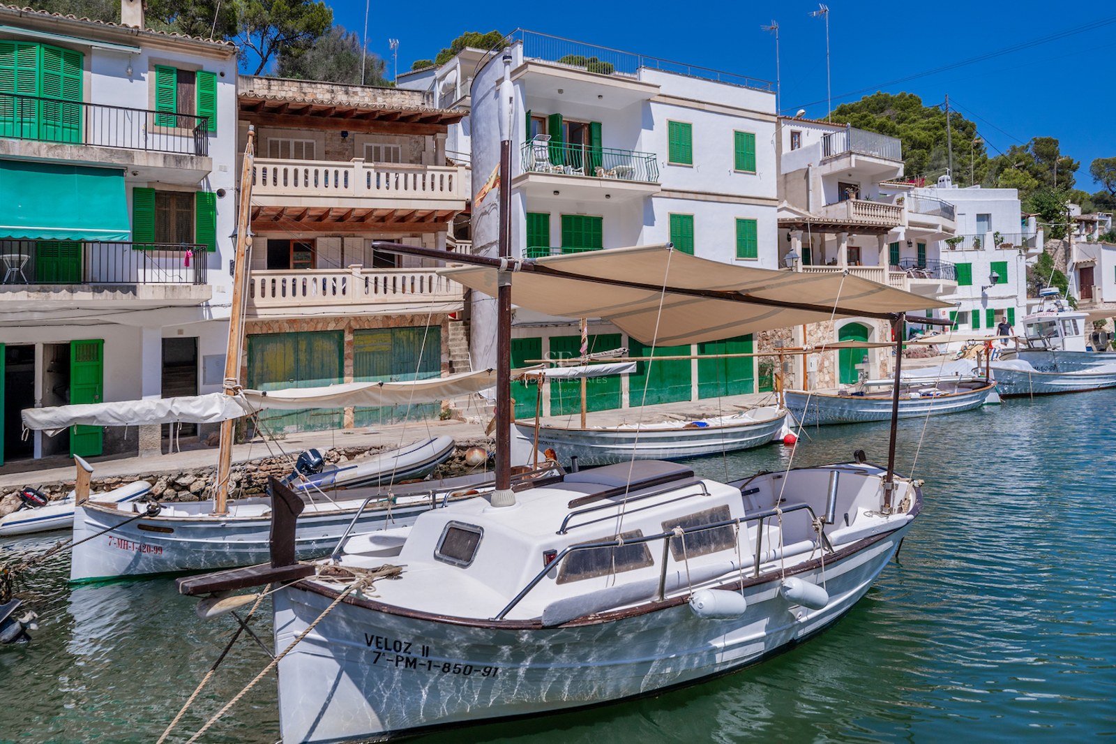 Traditional white houses with green shutters overlooking a calm harbor with classic wooden boats on turquoise water.
